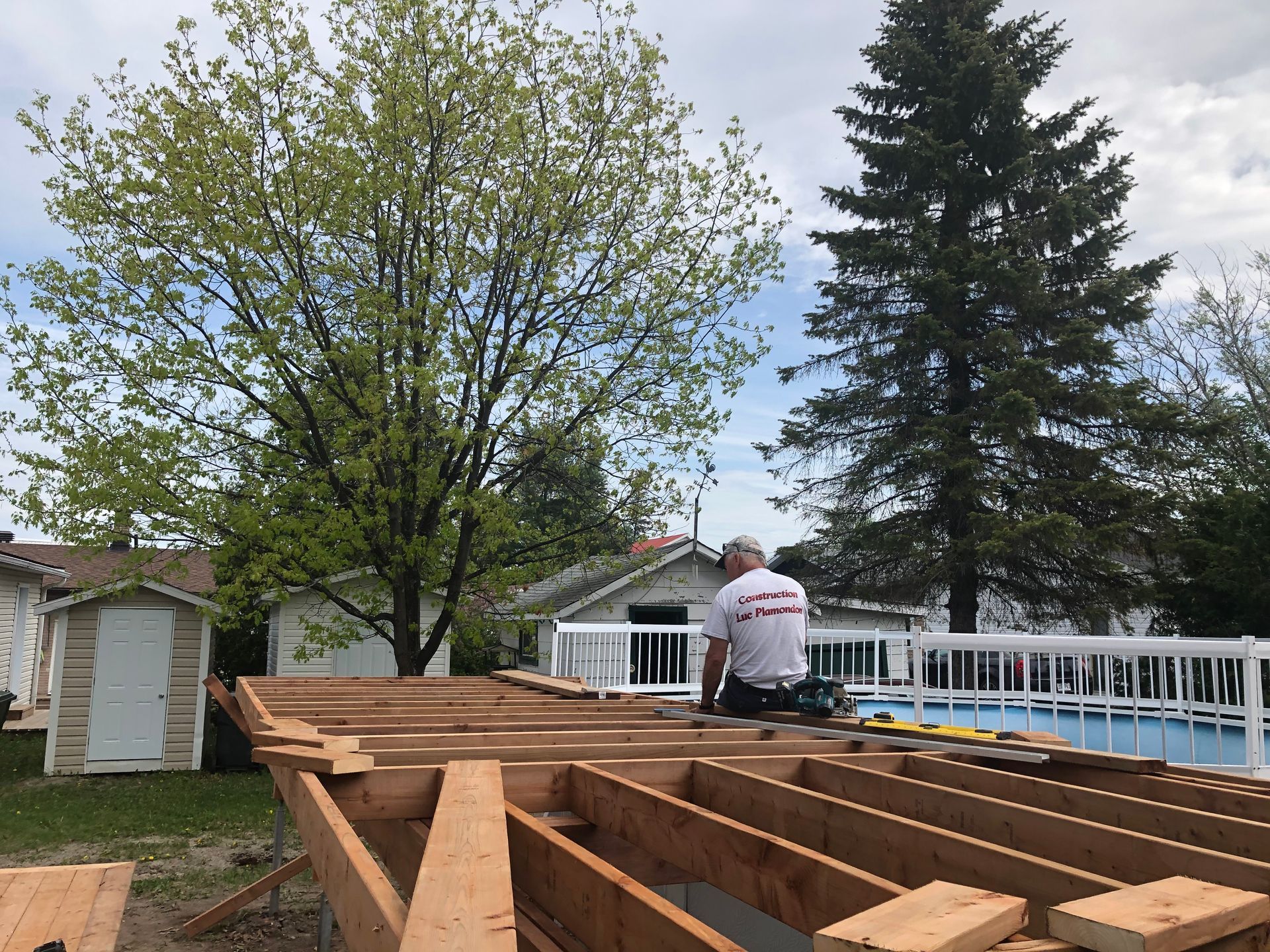 Un homme travaille sur la charpente d'une terrasse près d'une piscine. Un abri de jardin se dresse à l'arrière-plan.
