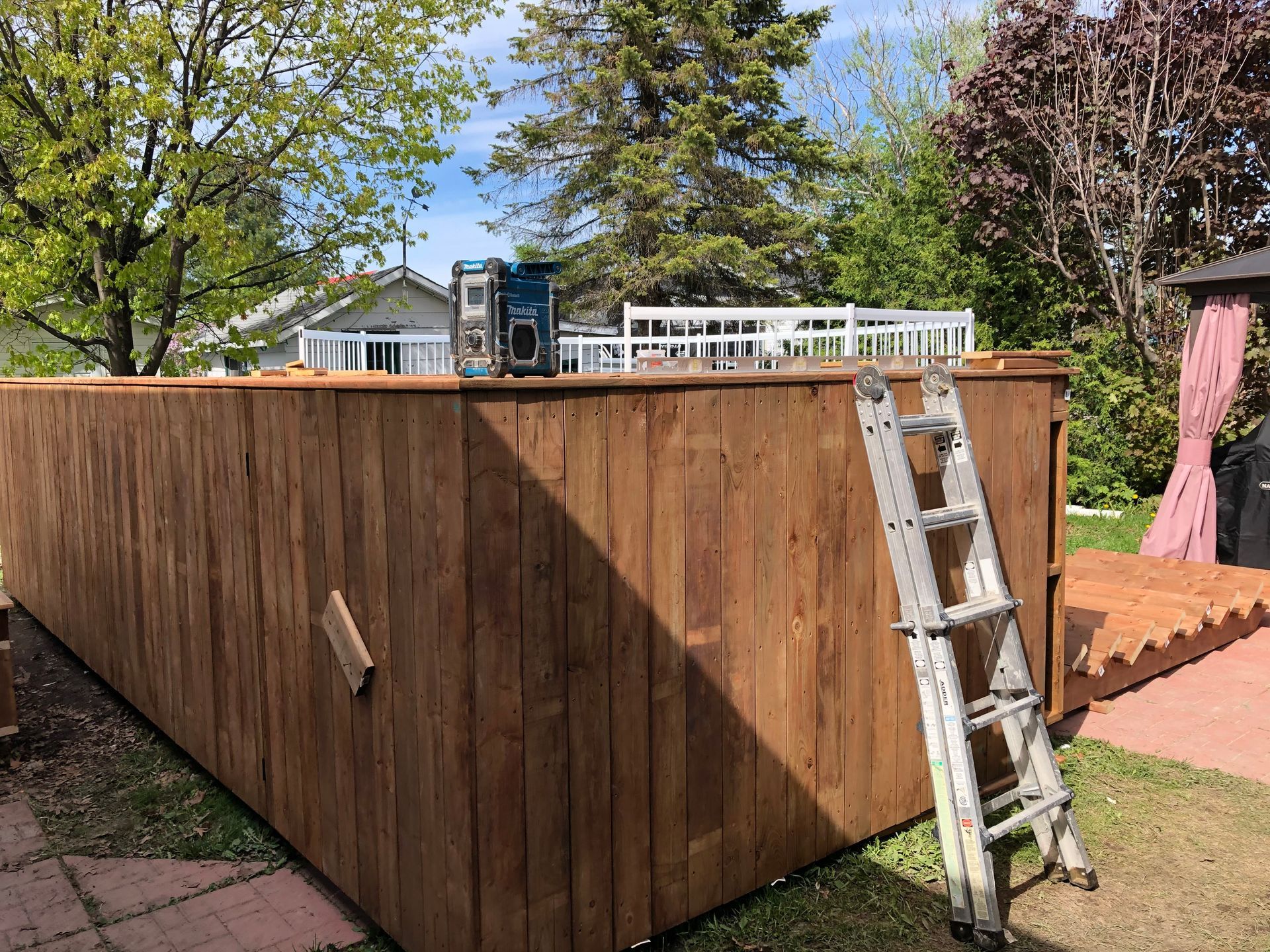 Une clôture en bois est en construction dans un jardin, une échelle est appuyée contre celle-ci.
