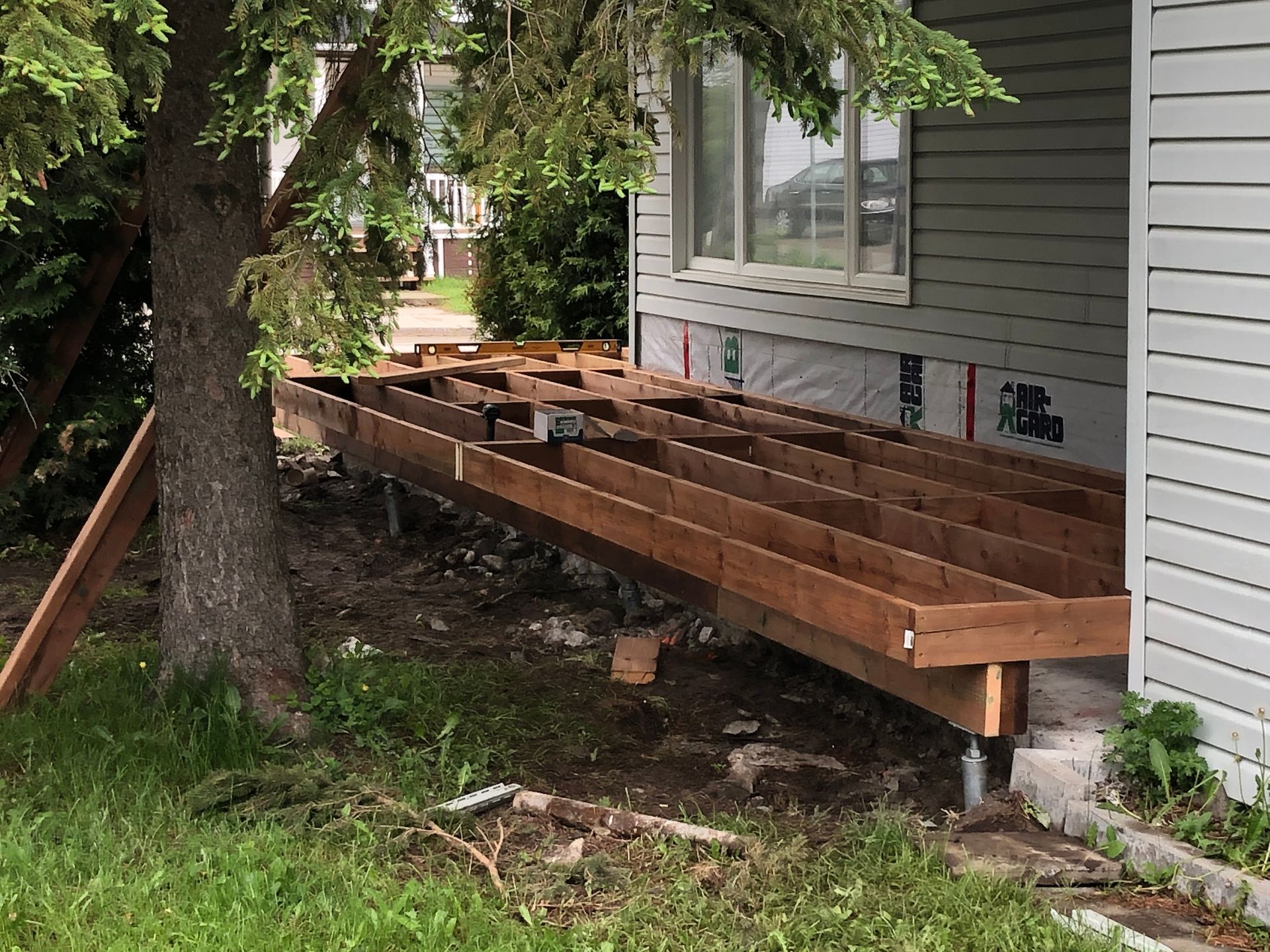 Terrasse en construction à côté d'une maison et d'un arbre. Poutres et charpente en bois brun.