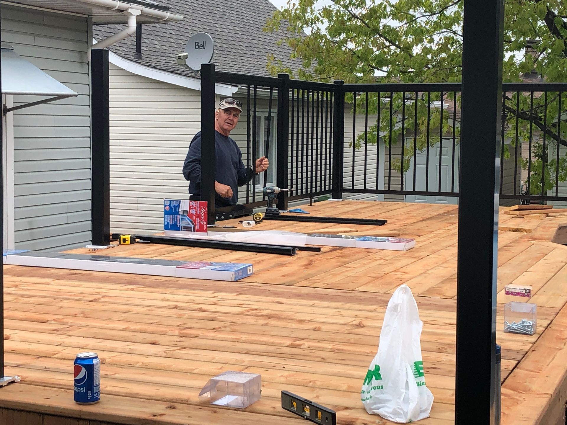 Un homme installe une rambarde noire sur une terrasse en bois. Outils et matériaux sont éparpillés.