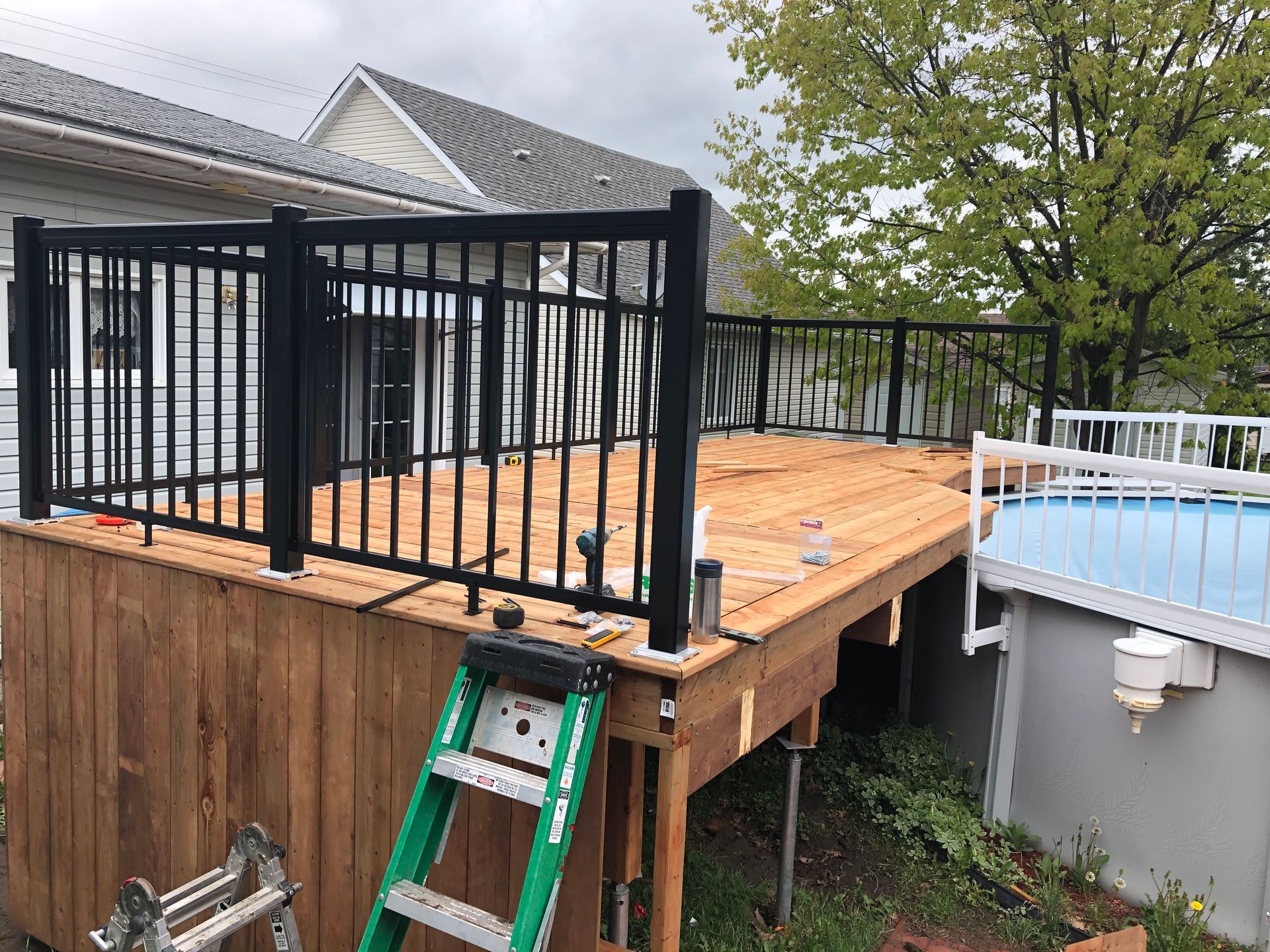 Une terrasse en bois nouvellement construite avec une rambarde en métal noir, près d'une maison et d'une piscine hors sol.