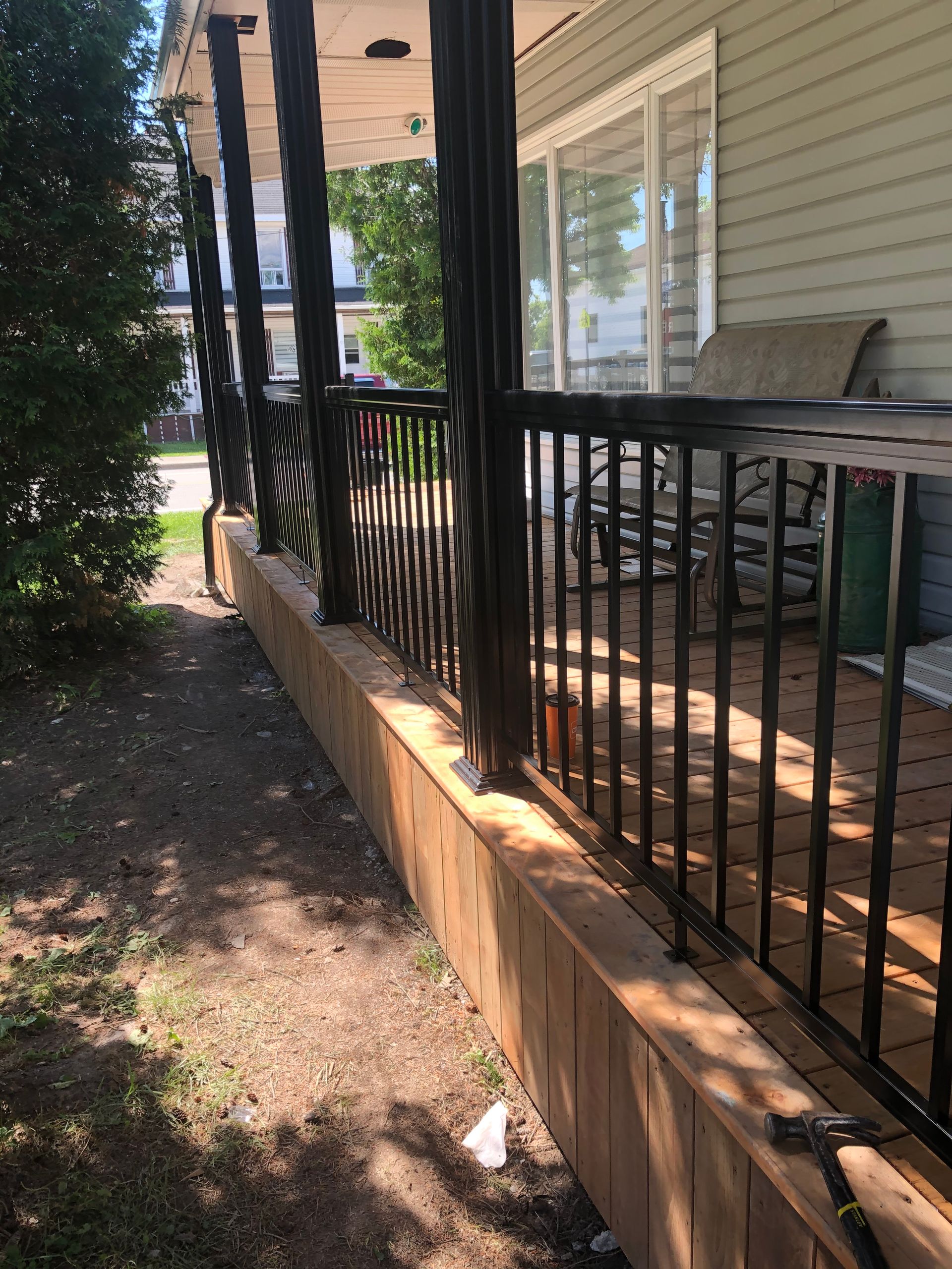 Une véranda avec des balustrades noires, une terrasse en bois et des colonnes de soutien ; une cour gazonnée sur la gauche.