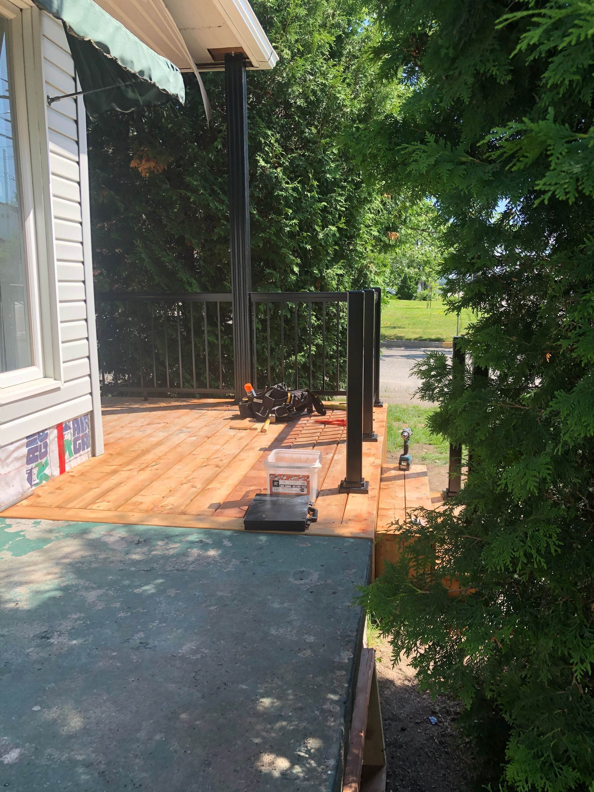 Terrasse en bois avec rambarde et poteaux noirs ; seau blanc et balance sur la terrasse. Feuillage vert à droite.