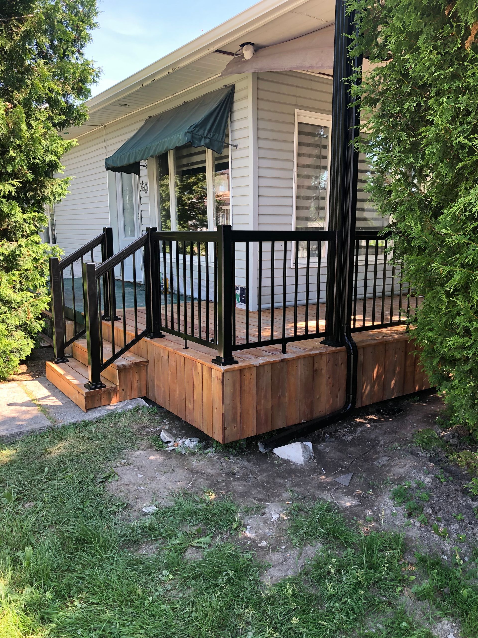 Terrasse en bois avec rambarde et marches noires devant une maison grise avec un auvent vert.