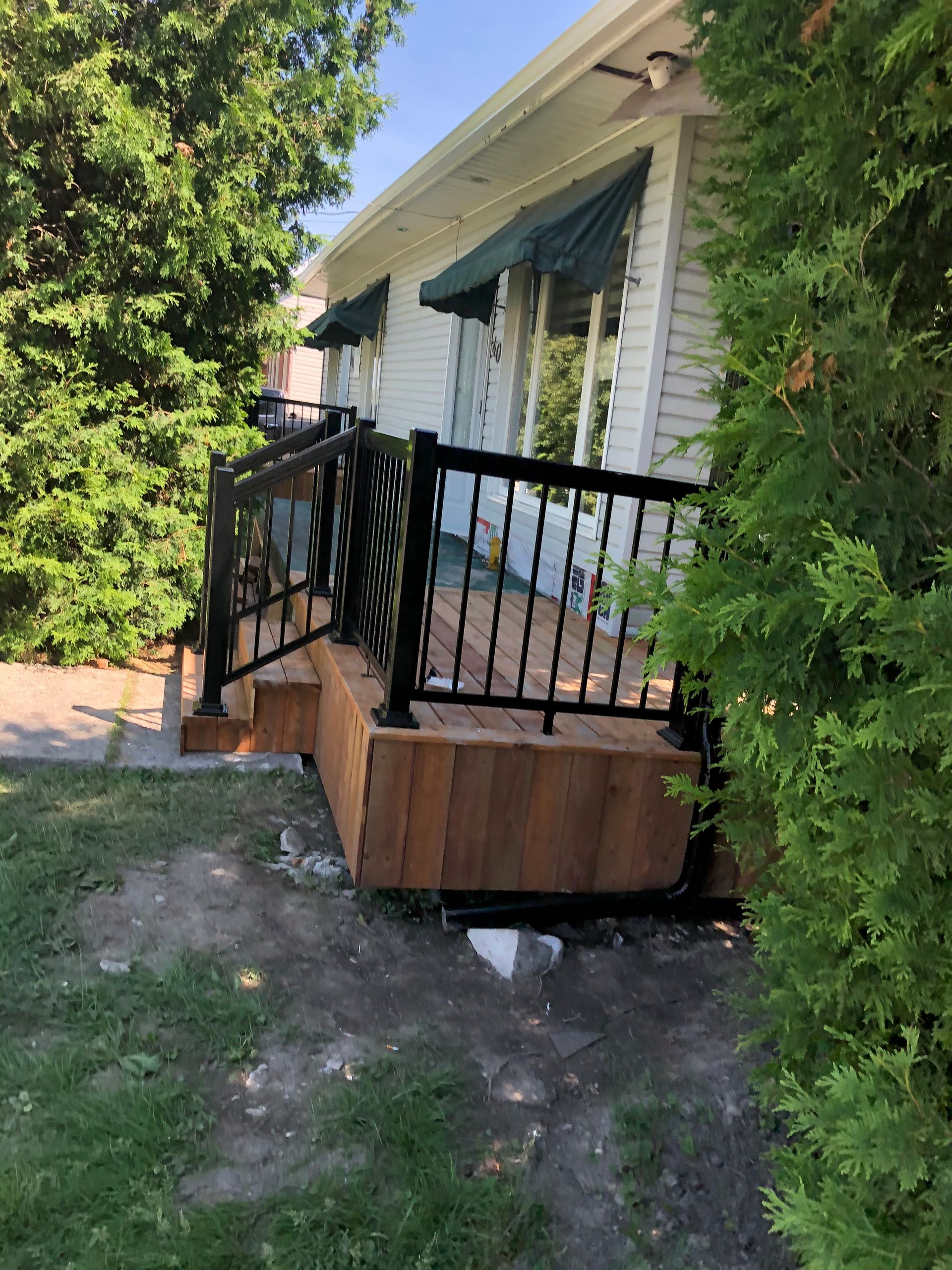 Terrasse en bois avec rambarde noire devant une maison blanche aux auvents verts, entourée d'arbres et de pelouse.