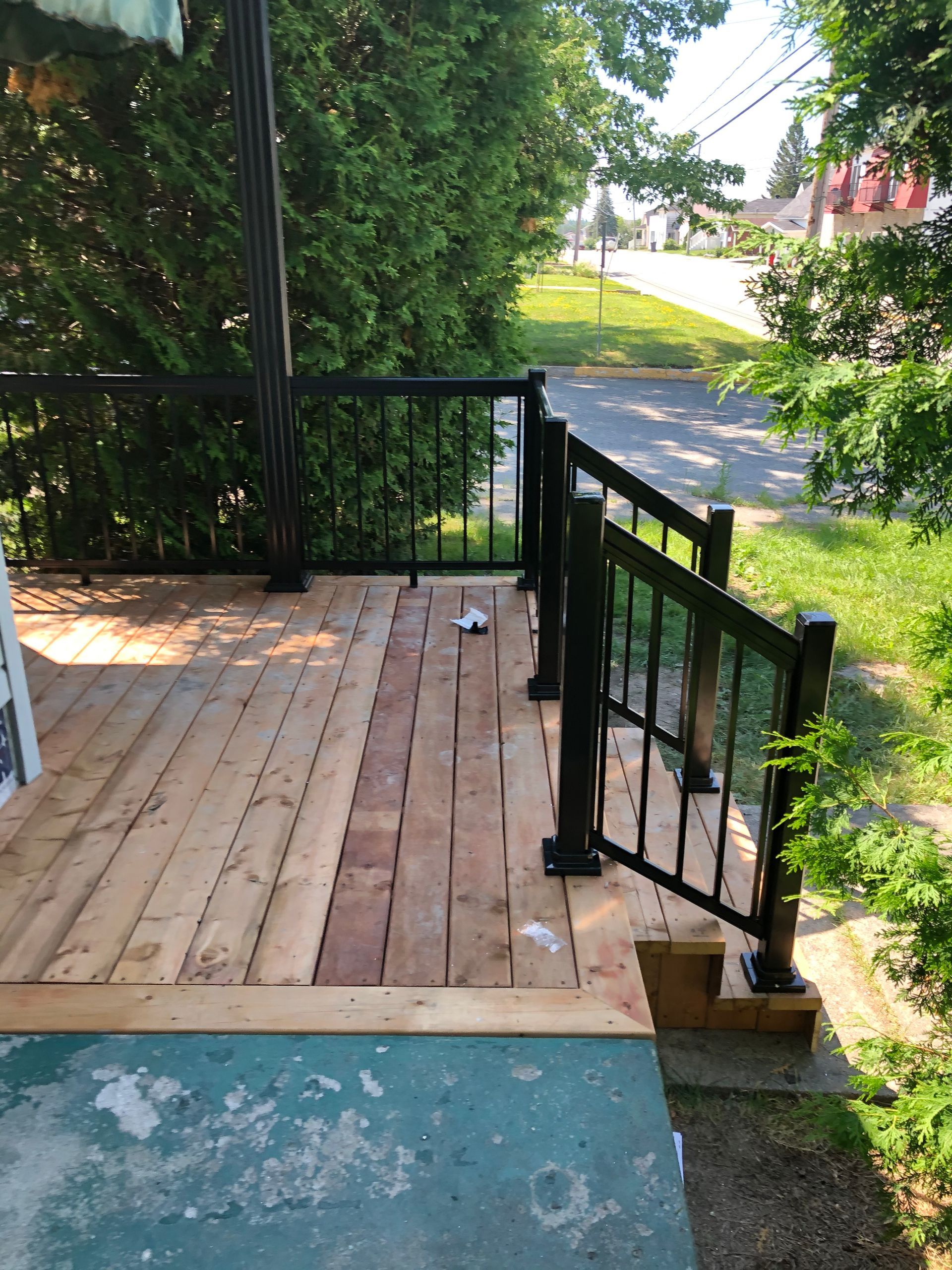 Terrasse en bois avec rambarde noire. Rue et arbres en arrière-plan.