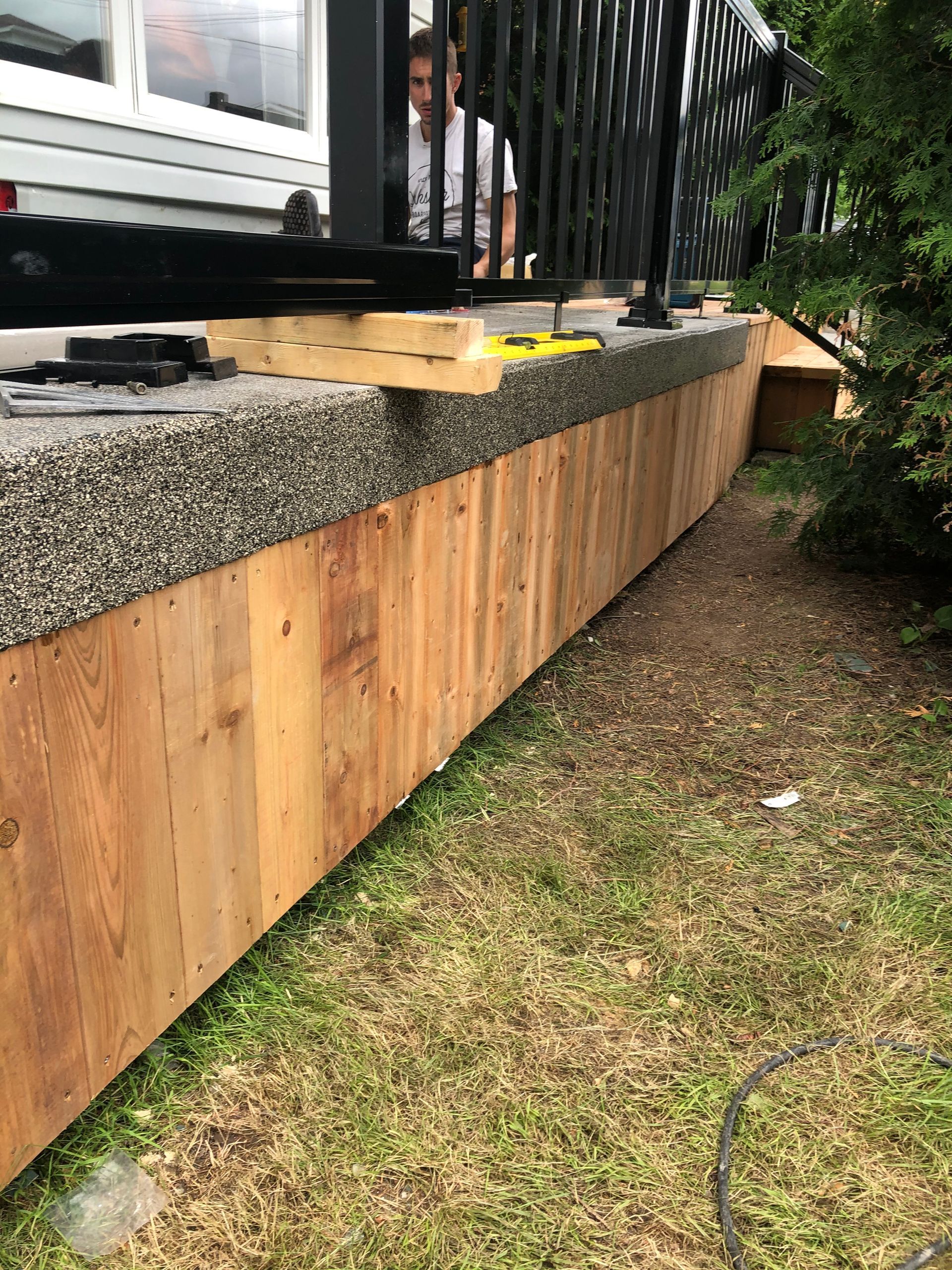 On installe un bardage en bois sur un mur de soutènement incurvé, sous une terrasse avec rambarde. Une personne est visible.