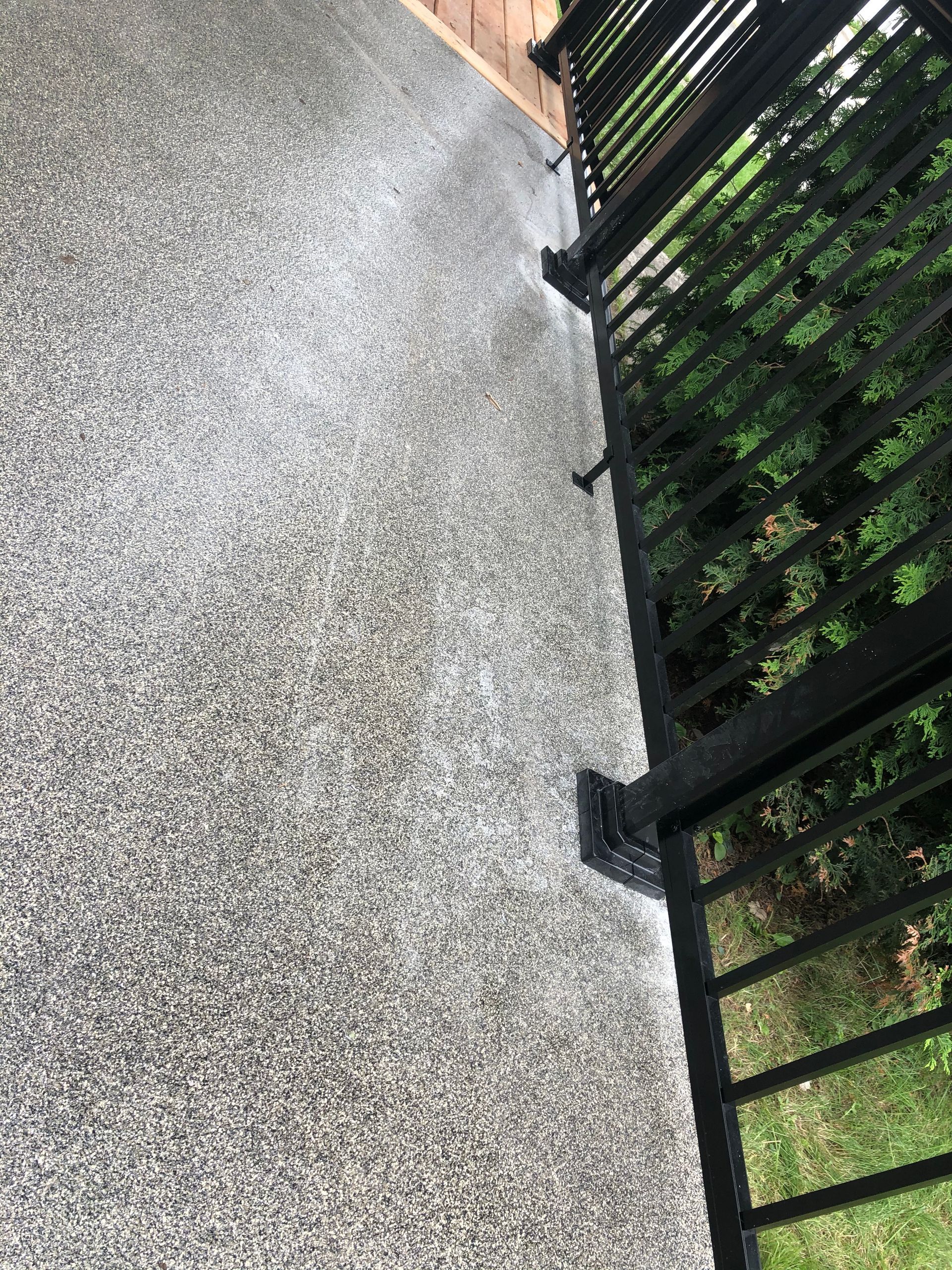 Terrasse en béton gris texturé avec rambarde en métal noir à droite, feuillage vert en arrière-plan.