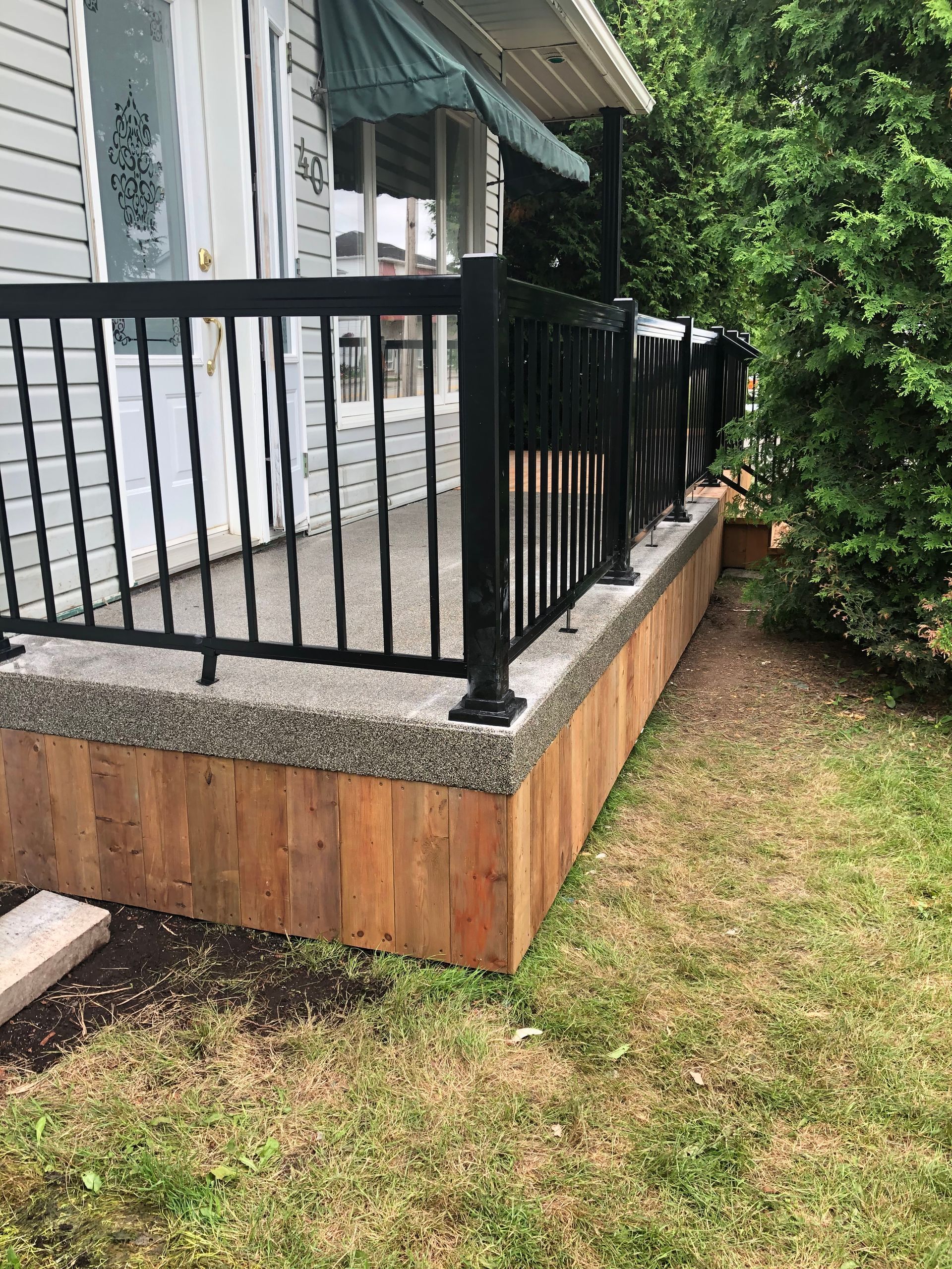 Un porche d'entrée avec une balustrade noire, du béton clair et des panneaux de bois. Il est entouré de pelouse verte.