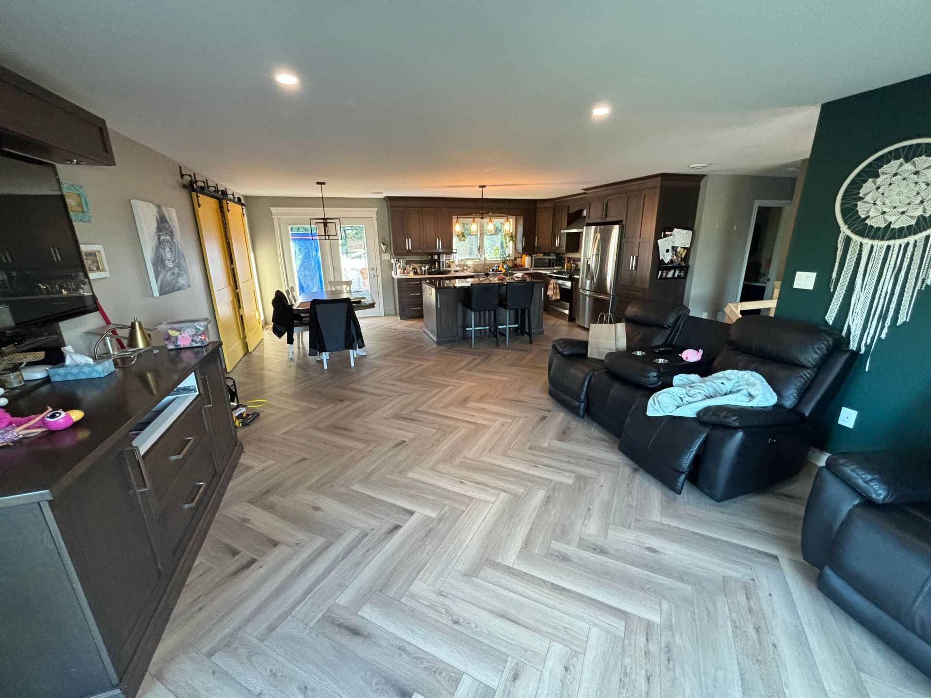 Espace de vie ouvert avec parquet à chevrons, cuisine et fauteuil inclinable noir. Placards, mur d'accent vert et télévision.