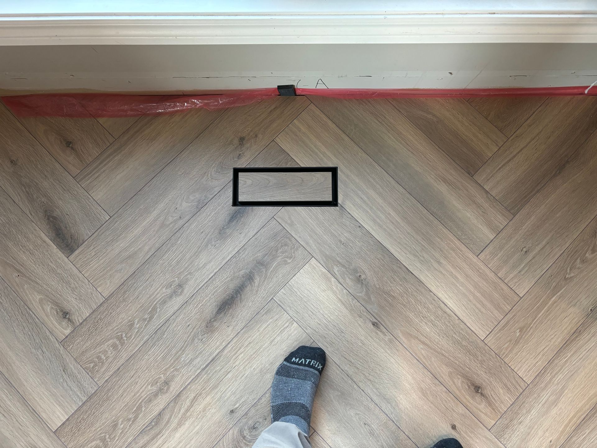 Parquet à chevrons avec une grille d'aération rectangulaire noire et le pied d'une personne portant une chaussette grise.