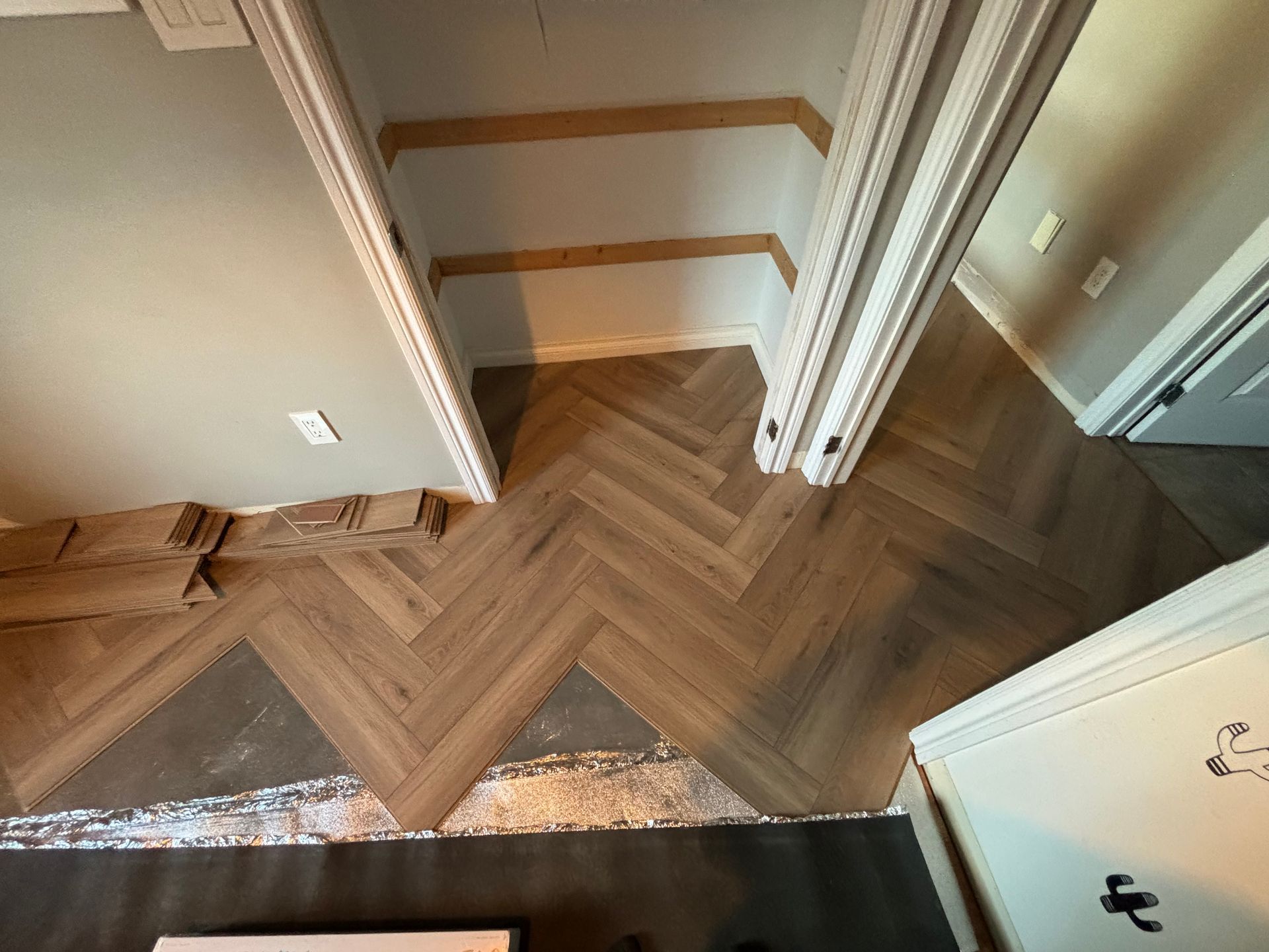 Pose de parquet à chevrons en cours, lames brun clair, encadrement de porte, moulures blanches, mur gris.