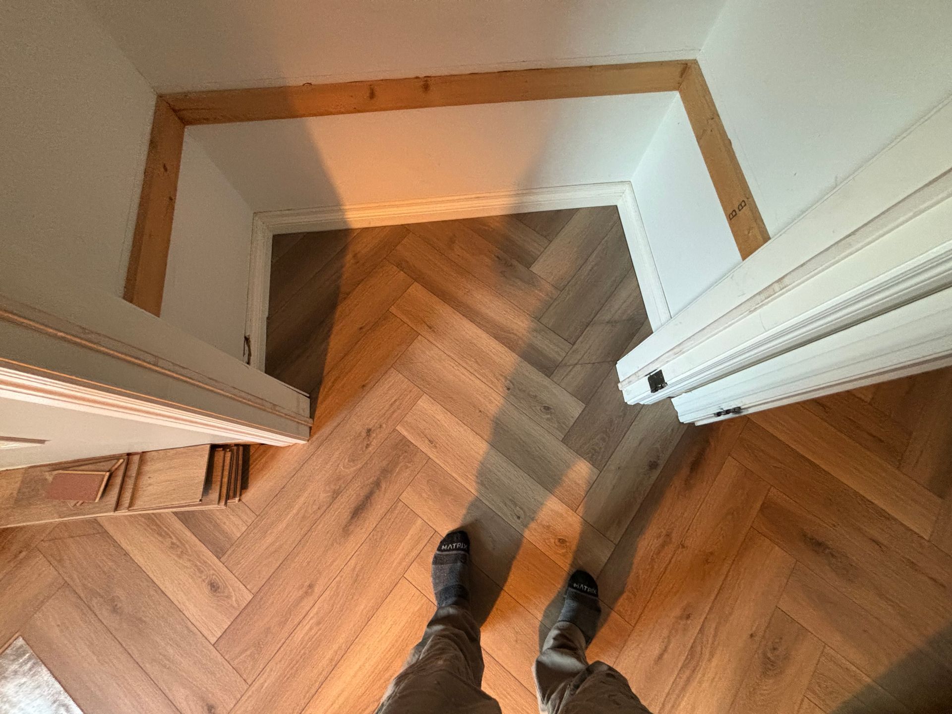 Parquet à chevrons dans l'encadrement de la porte, tons bruns et beiges. Boiseries, murs blancs. On aperçoit des pieds.