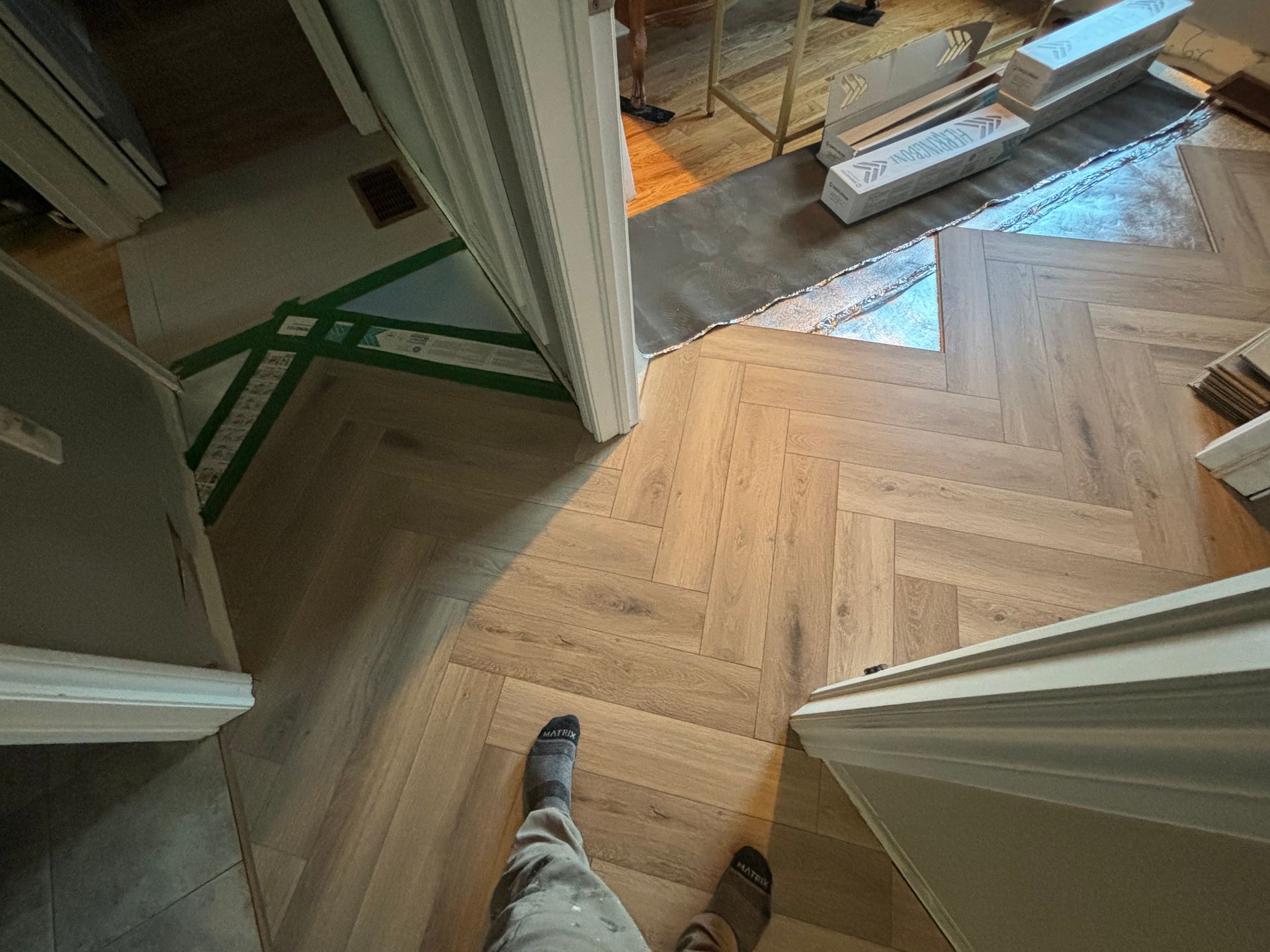 Pose d'un parquet clair à chevrons, laissant apparaître les jambes et les pieds d'une personne.