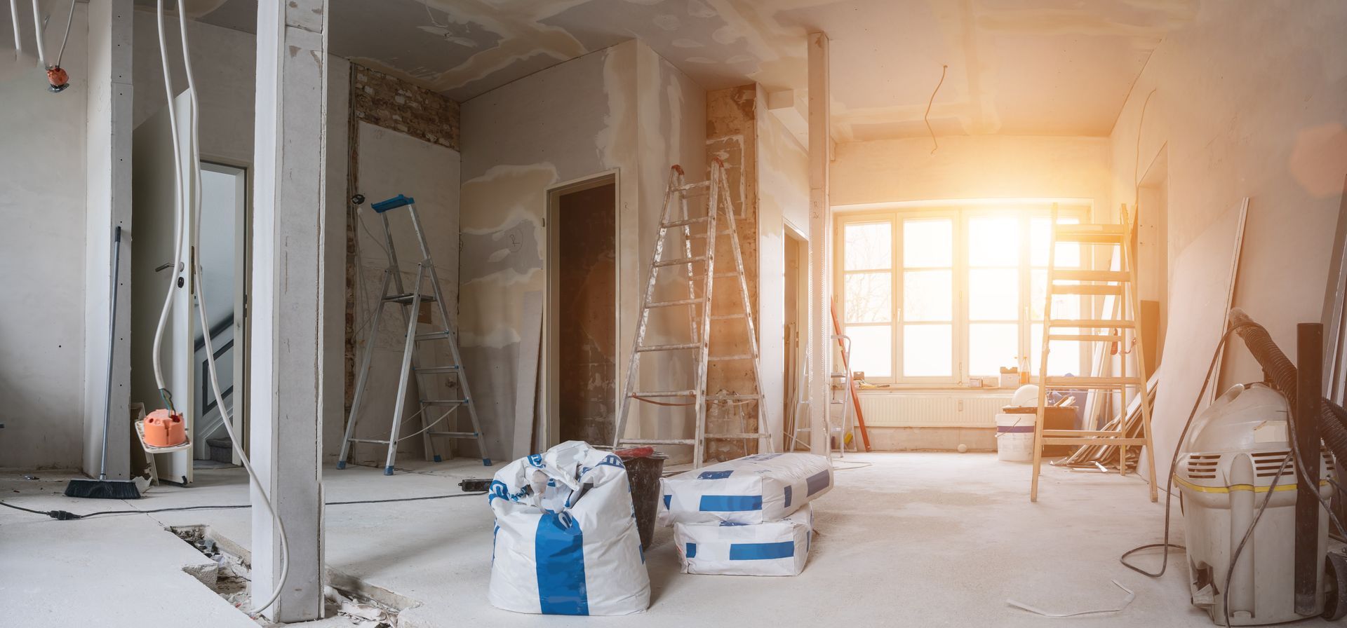 Pièce en rénovation ; échelles, sacs de matériaux et lumière vive du soleil entrant par une fenêtre.