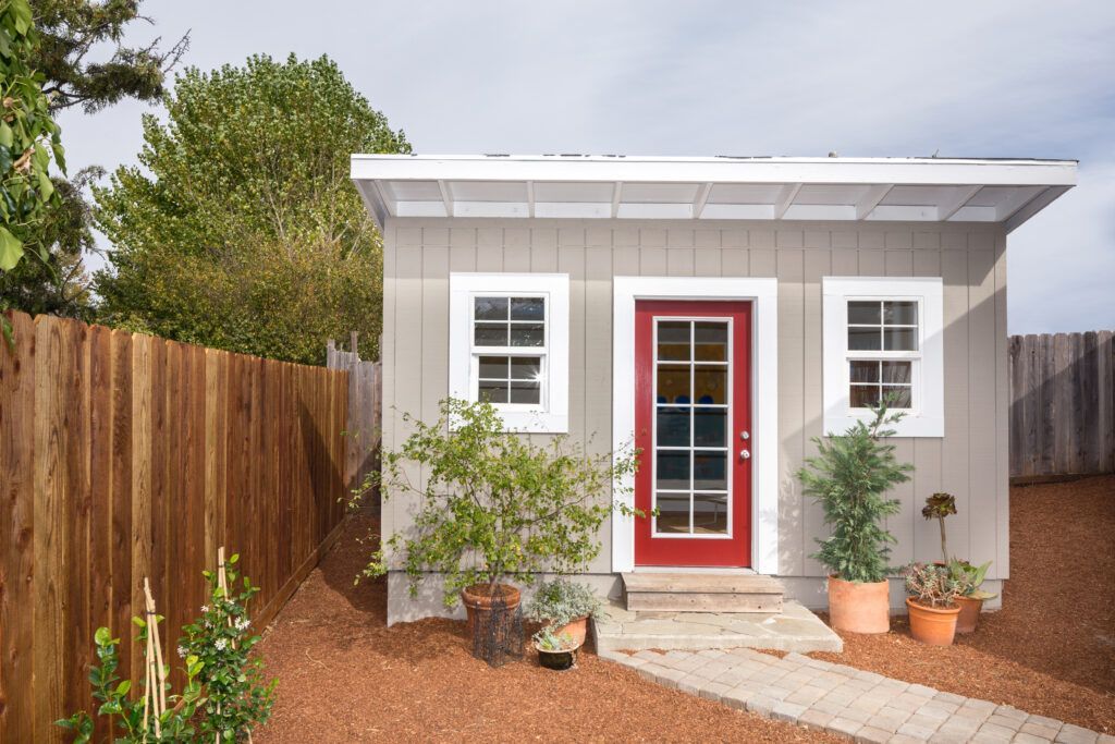Small gray cottage with a red front door, white-trimmed windows, and a desert-style front yard