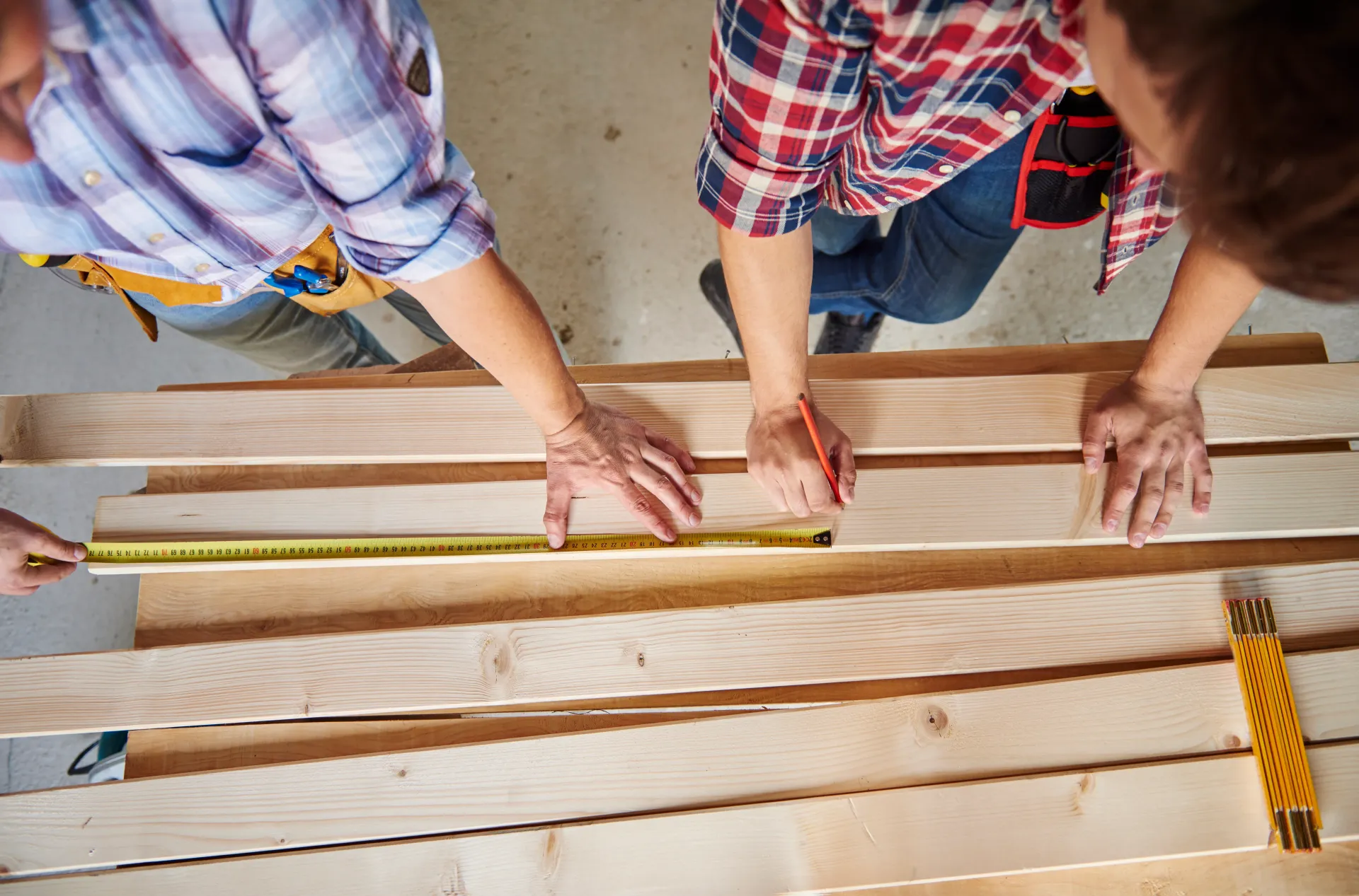 Three people measuring wooden boards with a tape measure at a construction site.