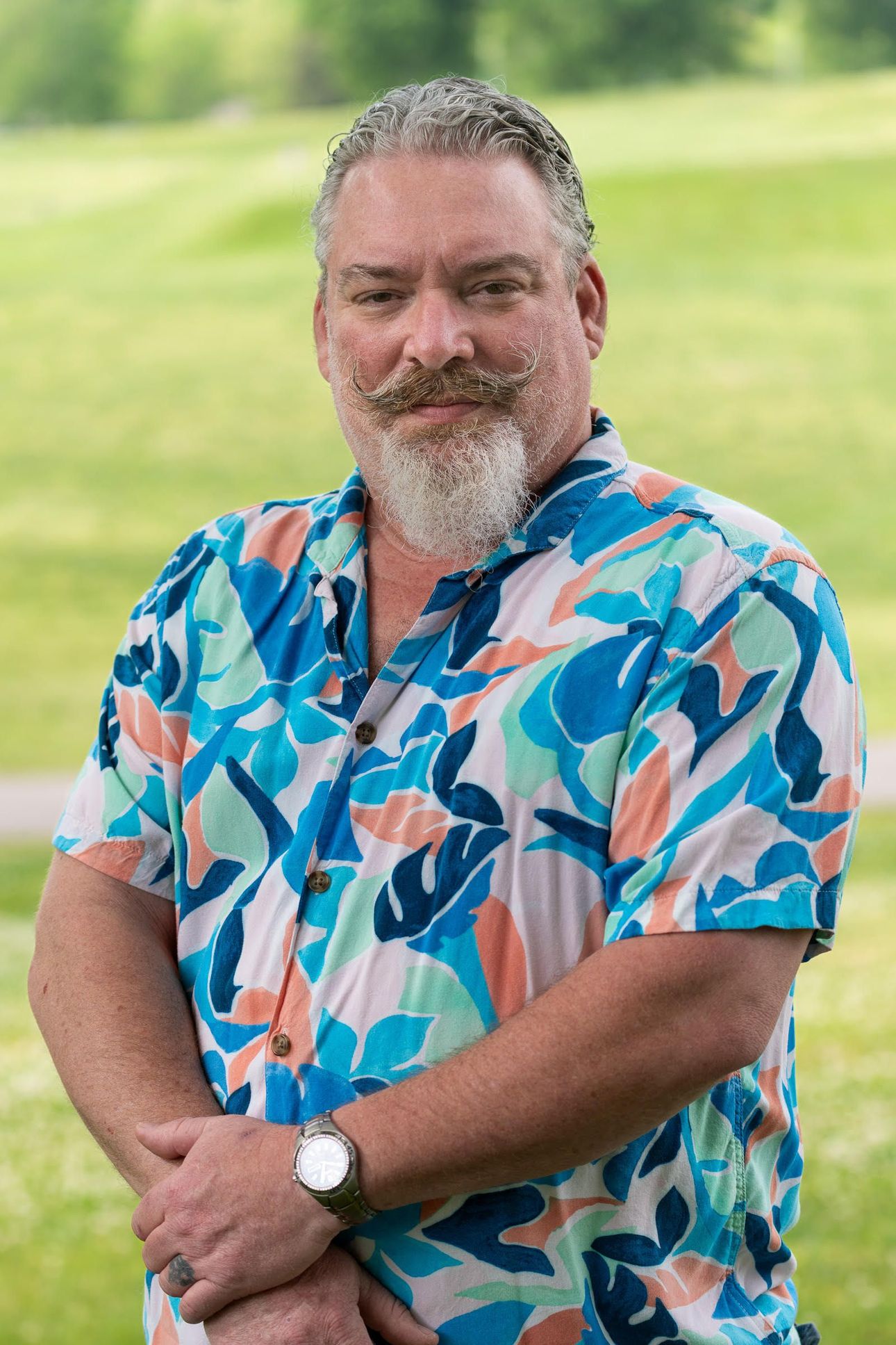 Smiling man in a colorful blue and pink shirt standing outdoors against a blurred green background