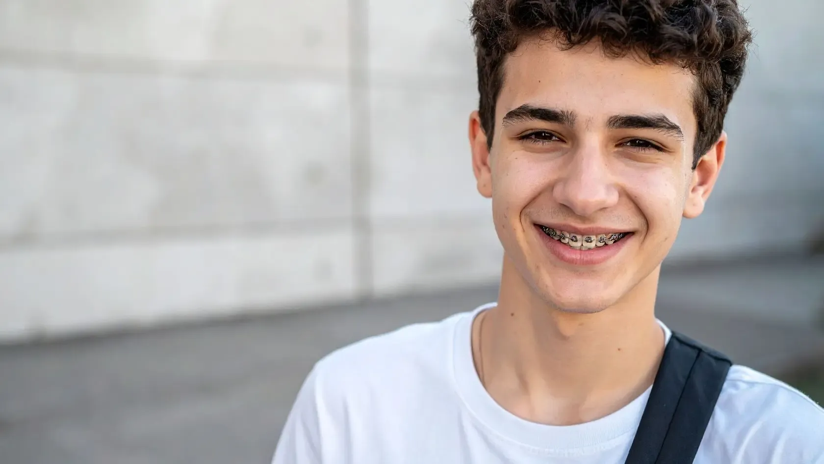 Teenager with braces smiles, wearing a white t-shirt and carrying a backpack. Outdoors, gray backdrop.