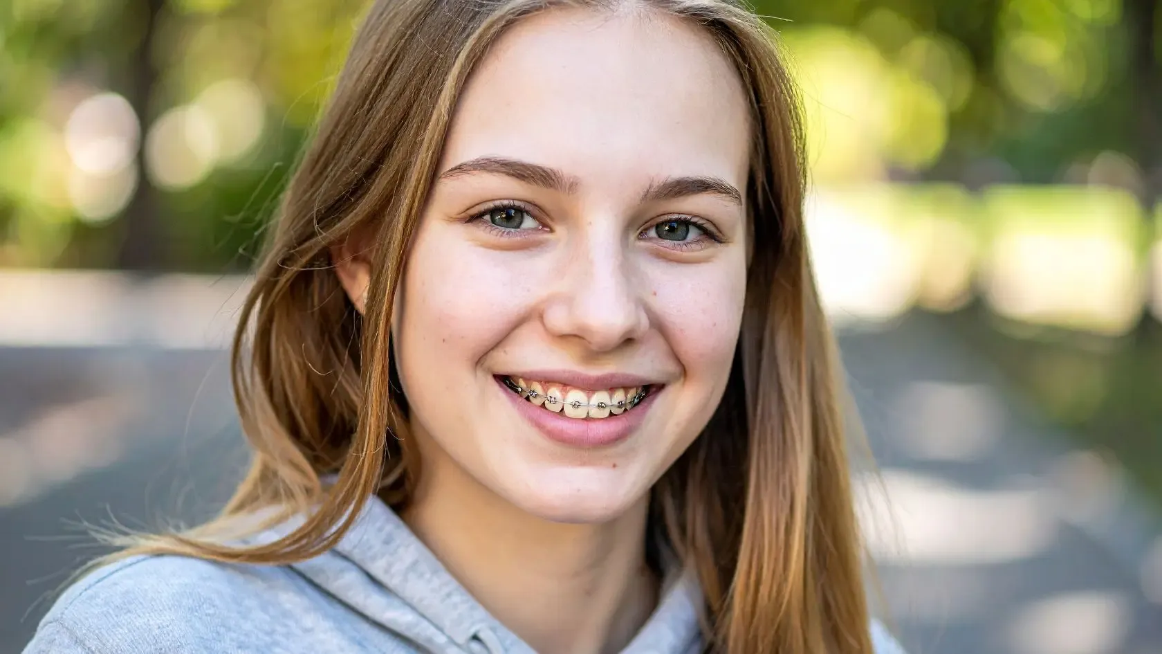 Smiling teenage girl with braces, long blonde hair, and a gray hoodie in an outdoor setting.