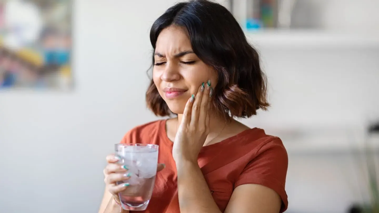 Woman holding glass of water, touching cheek in pain, likely from toothache.