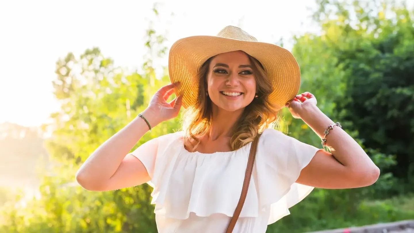 Woman in white top and straw hat smiles outdoors, holding hat with both hands.
