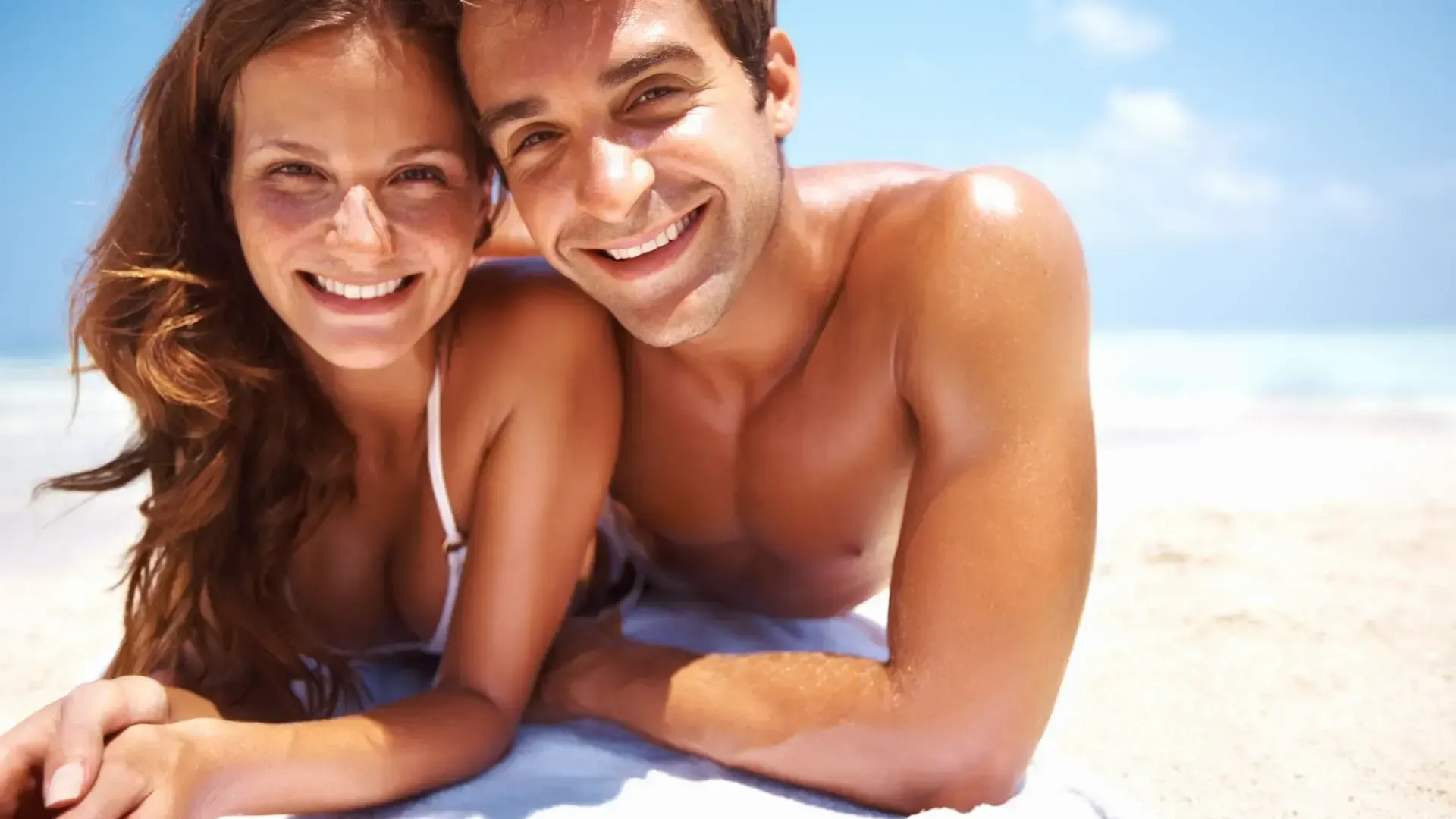 A man and a woman are laying on the beach and smiling for the camera.