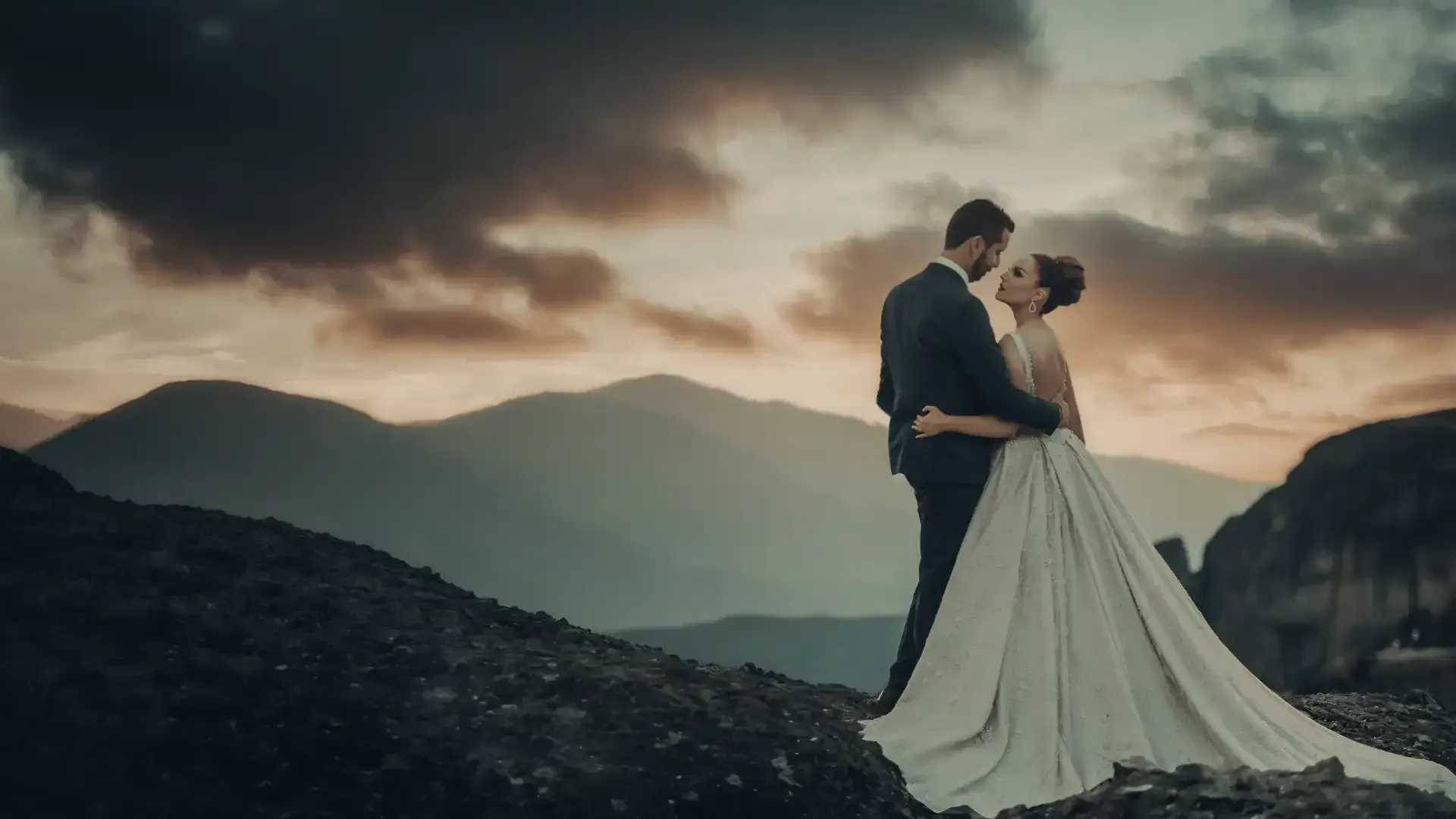 A bride and groom are standing on top of a mountain at sunset.