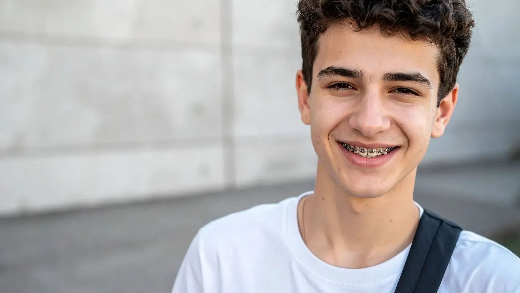 Teenager with braces smiles, wearing a white shirt and carrying a black bag, against a blurred gray wall.