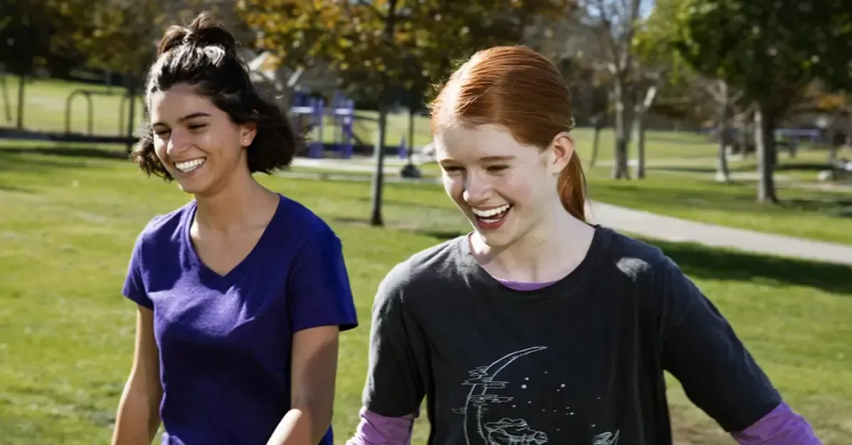 Two girls are walking in a park holding hands and smiling.