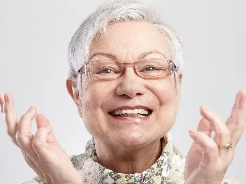 An elderly woman wearing glasses is smiling with her hands in the air.