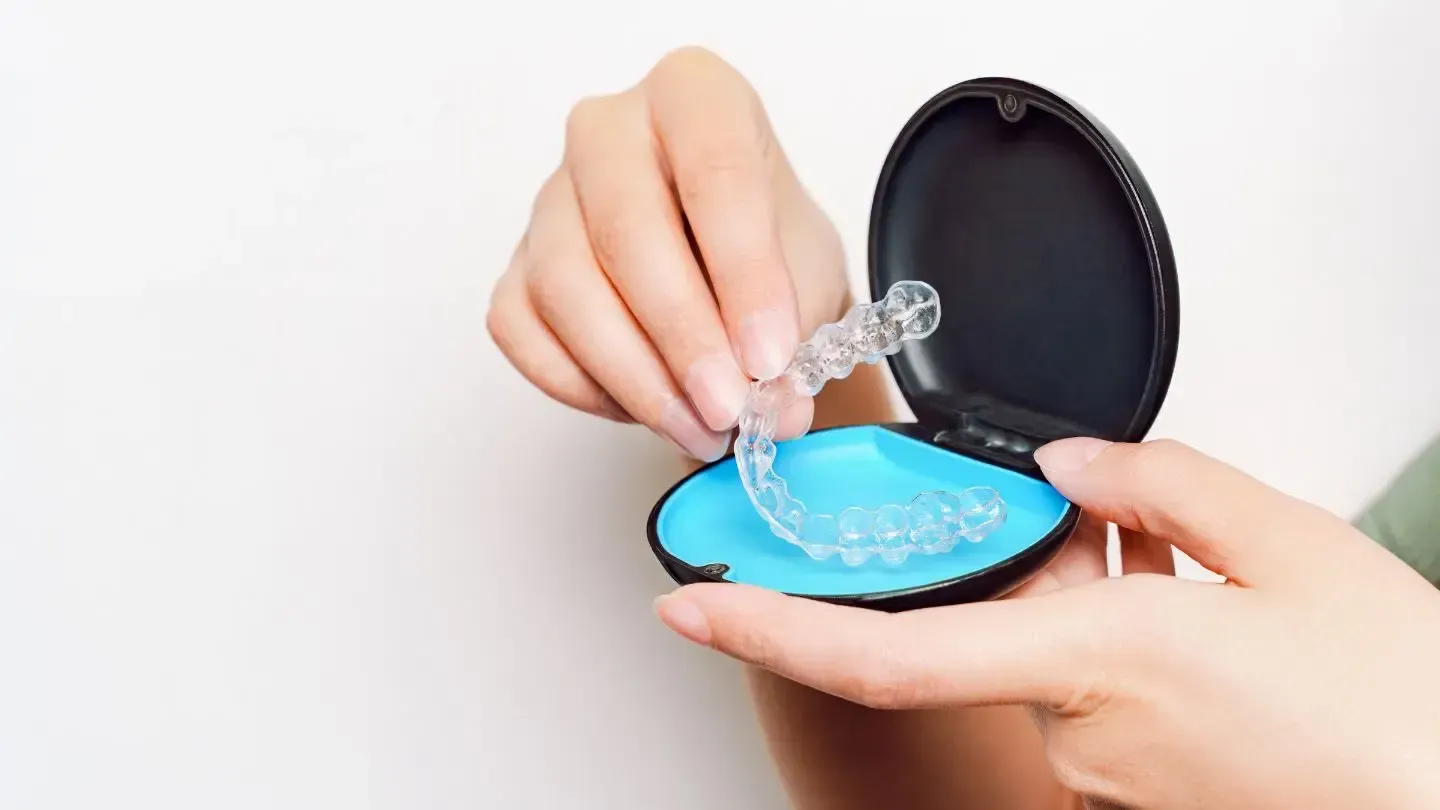 Hands holding a clear dental aligner over a blue and black carrying case.