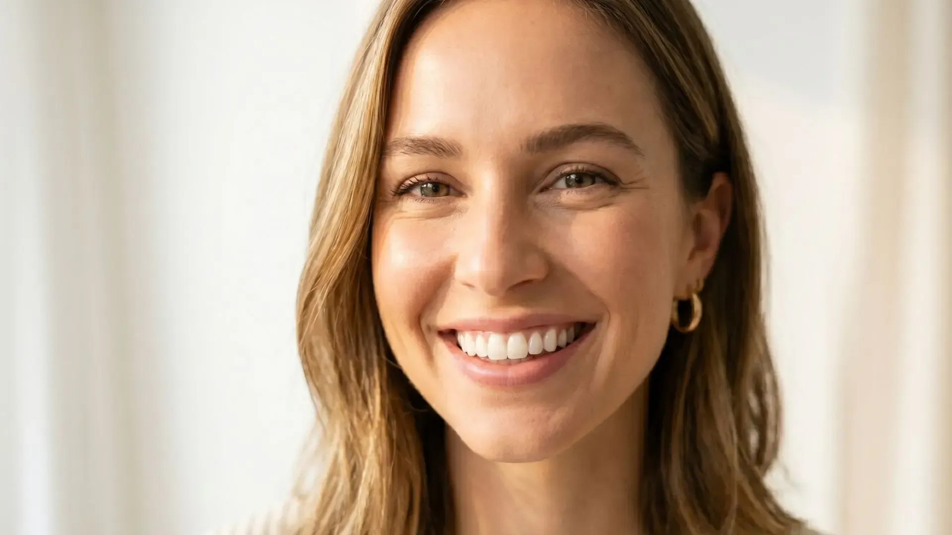 Smiling person with long brown hair, wearing gold hoop earrings, against a bright neutral background