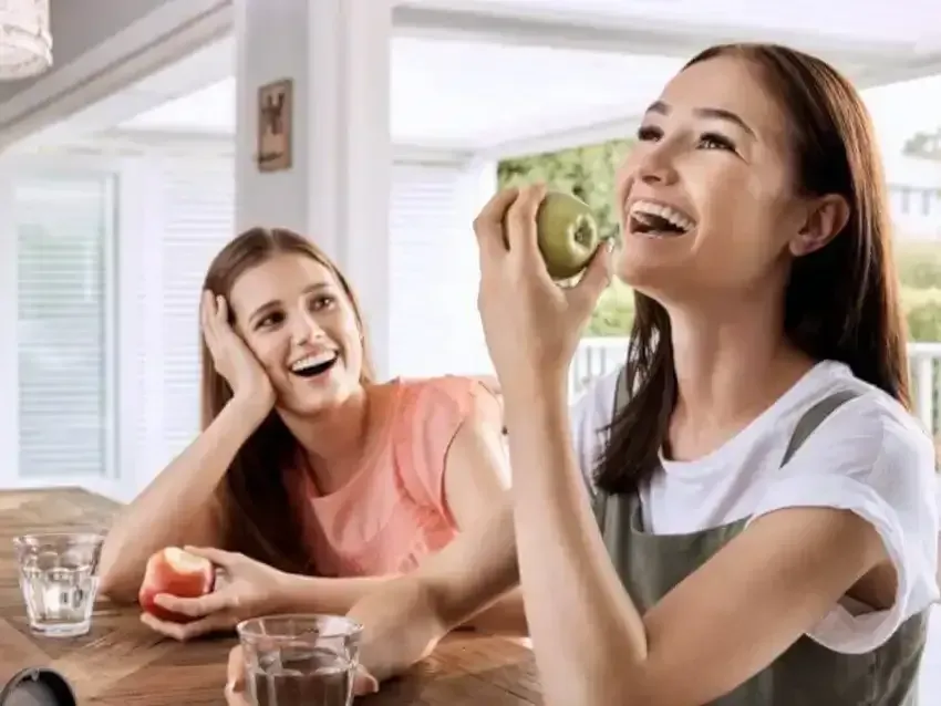 Two women are sitting at a table eating apples.