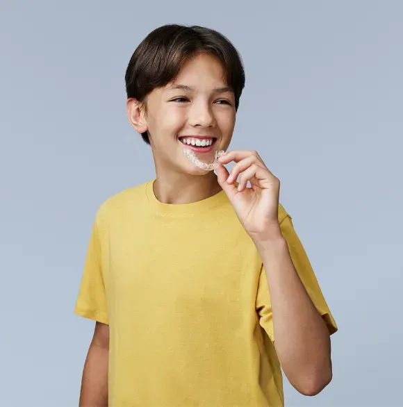 A young boy wearing a yellow shirt is smiling while holding a clear brace in his mouth.