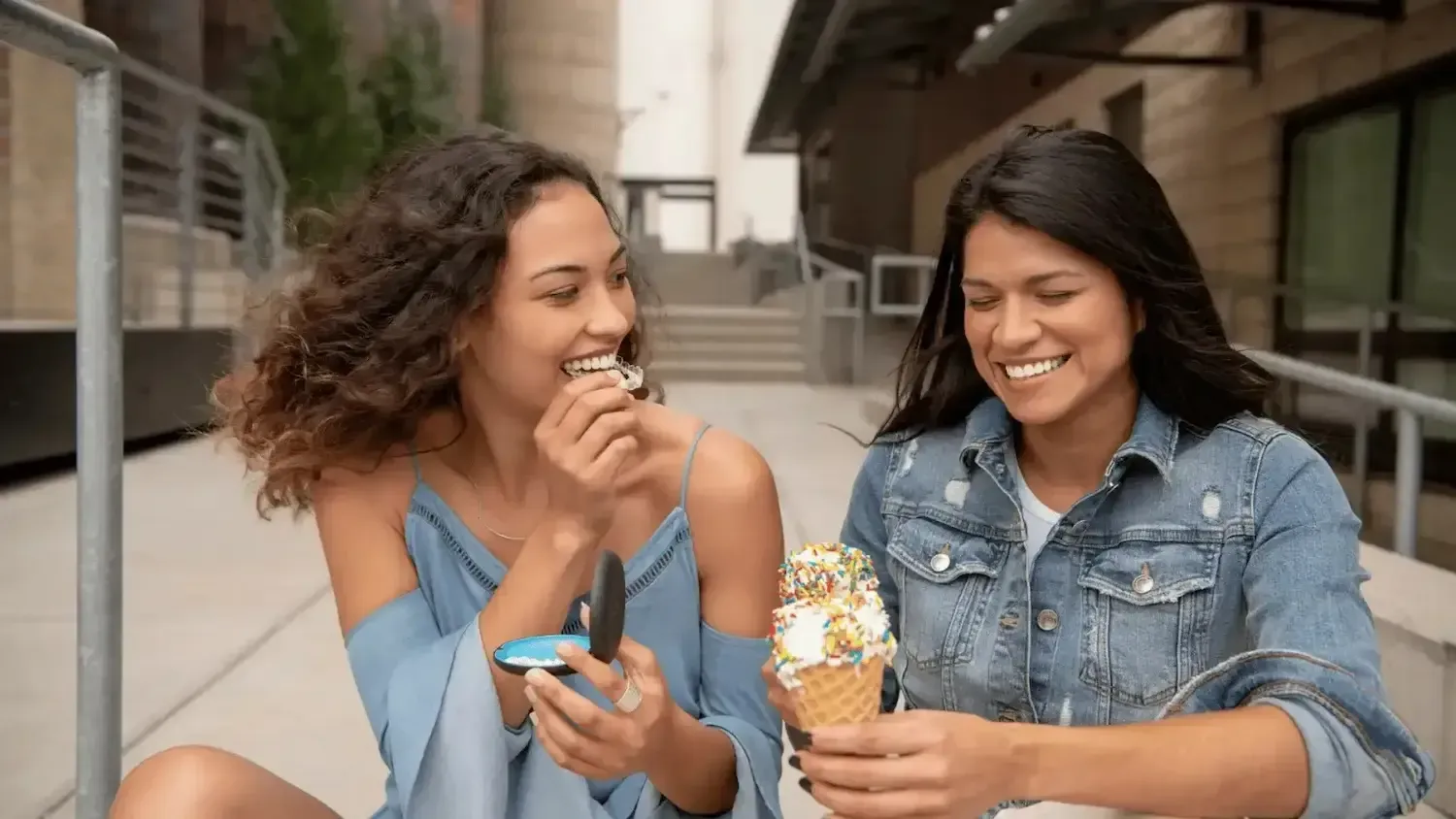 two women are sitting on a sidewalk eating ice cream cones .