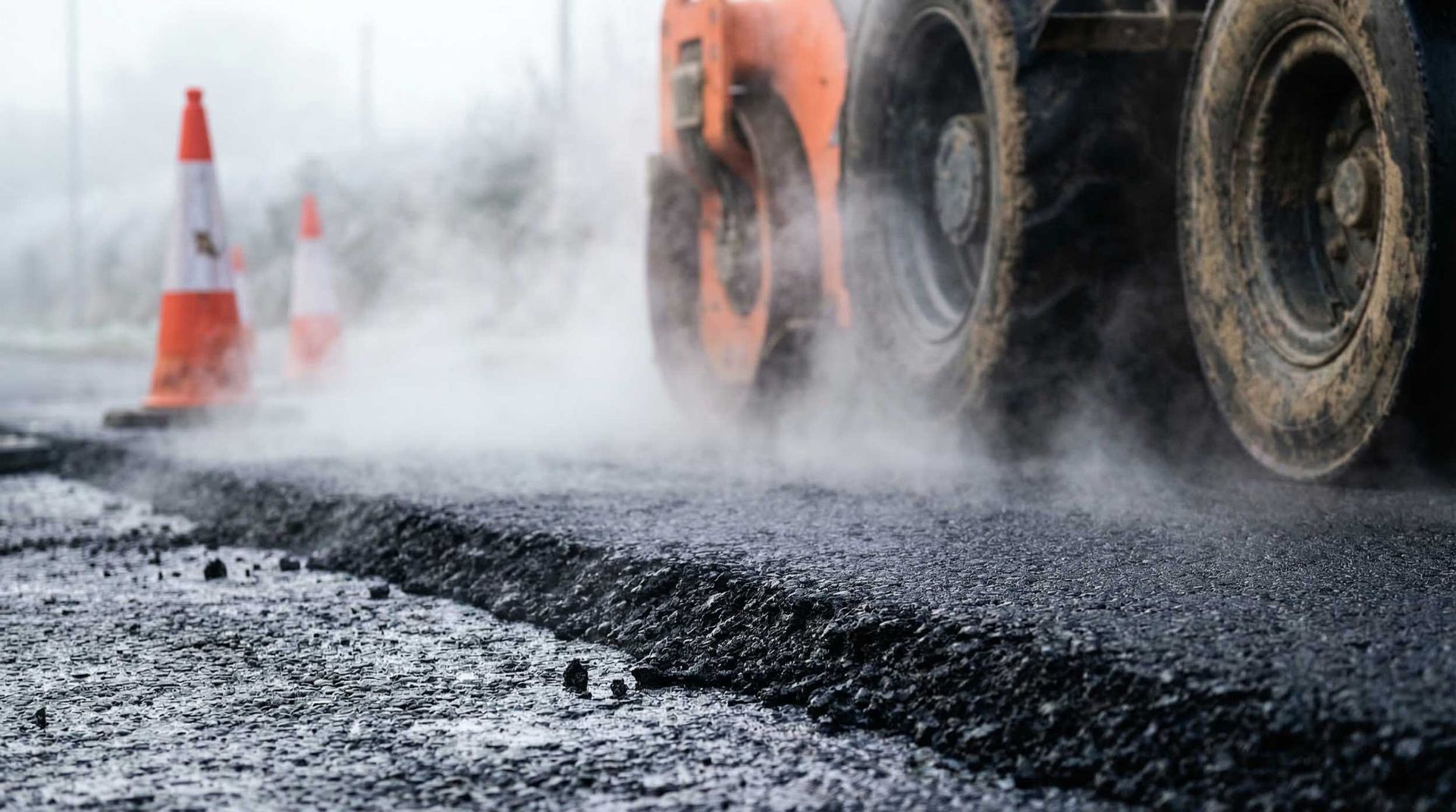 Steamroller paving wet asphalt beside an orange traffic cone, with steam rising over the road.