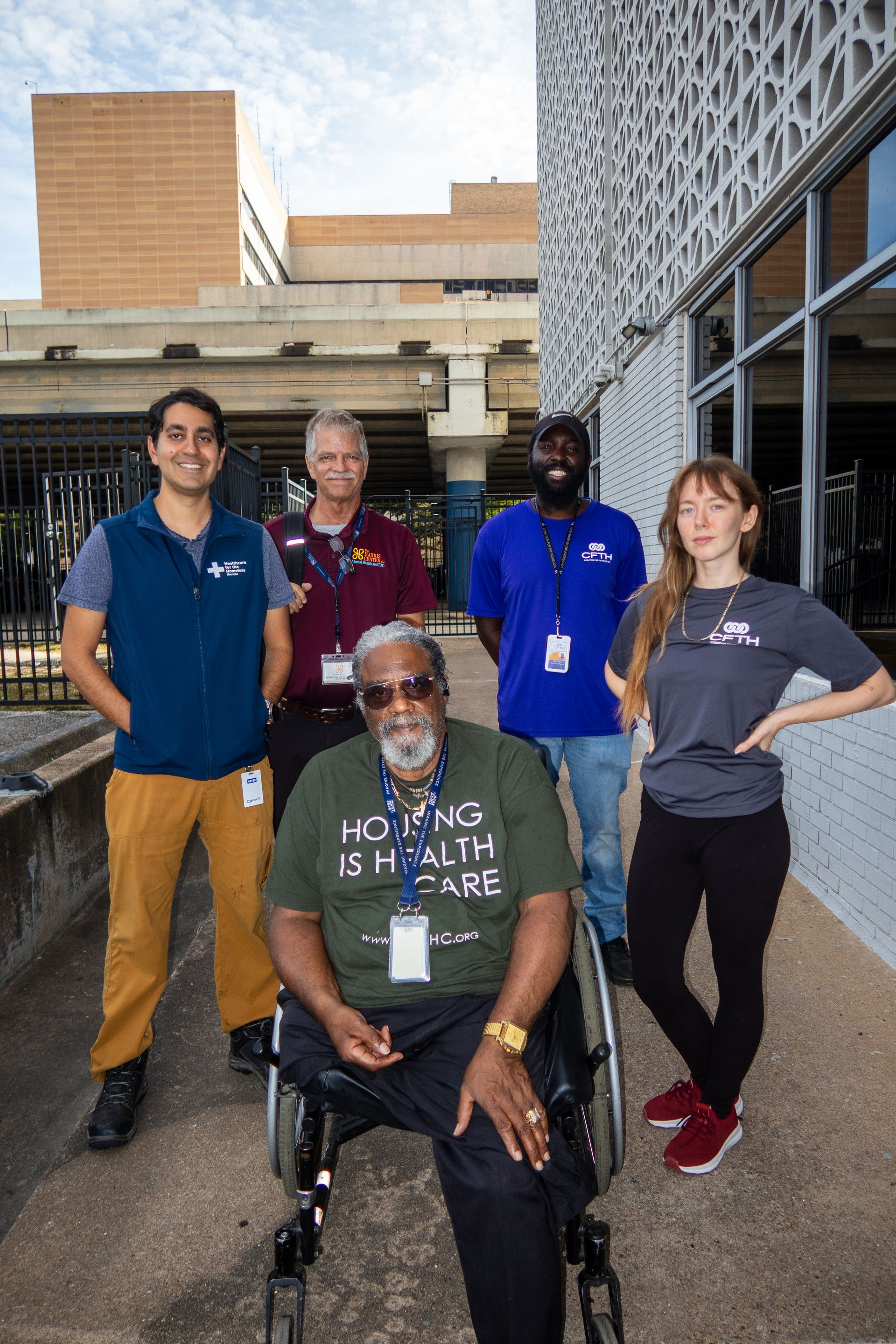 Five people pose outside a building; one seated in front, four standing behind, all smiling.