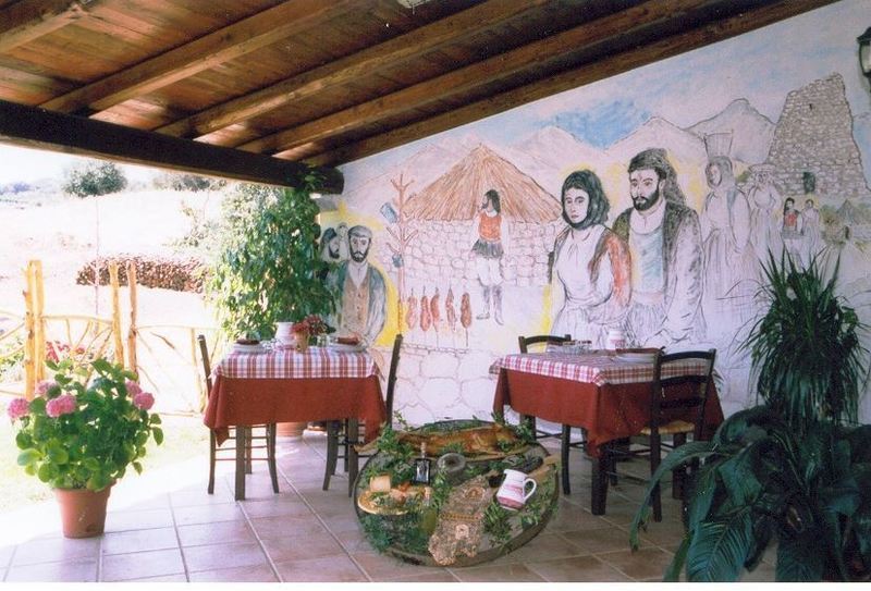 Una zona pranzo esterna coperta è arredata con due tavoli con tovaglie a quadri rossi, piante in vaso e un murale raffigurante delle figure.