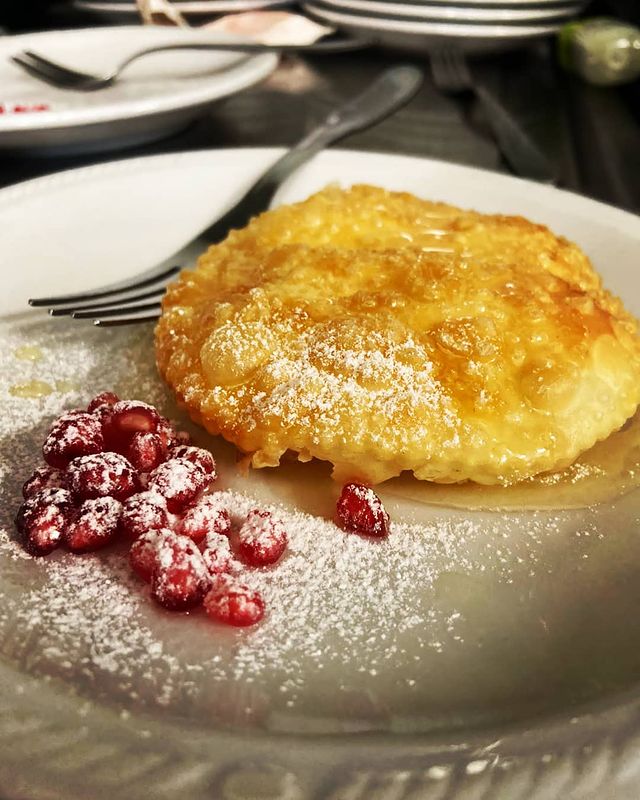 Una frittella dorata servita su un piatto, guarnita con sciroppo e zucchero a velo, accompagnata da chicchi di melograno freschi.