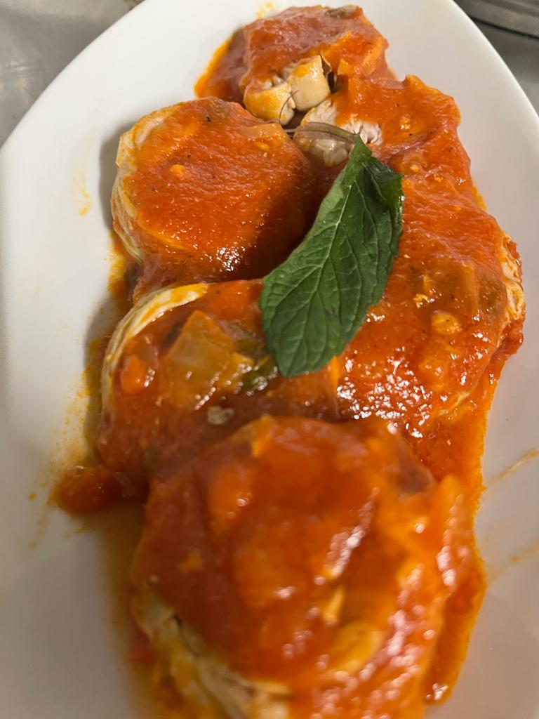 Tre filetti di pesce rotondi e cotti, ricoperti da una vivace salsa di pomodoro, disposti su un piatto ovale bianco e guarniti con una fogliolina di menta.