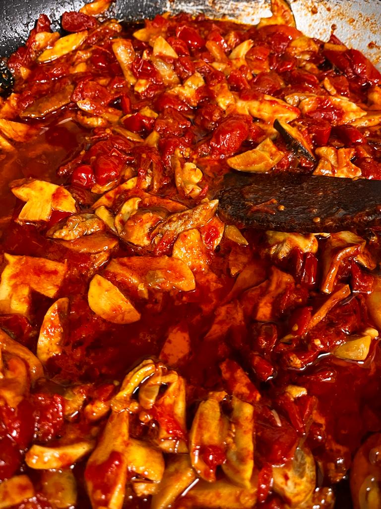 Un primo piano di funghi affettati che saltano in padella con una salsa di pomodoro di un rosso acceso e una spatola da cucina scura.