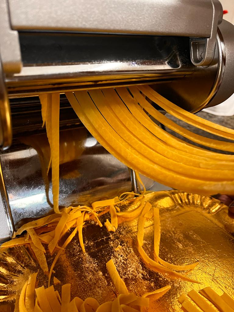 Una macchina per la pasta cromata estrude fettuccine fresche di colore giallo dorato su un vassoio dorato lucido sottostante.