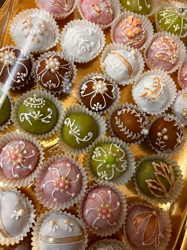 Una vista dall'alto di un assortimento di dolcetti colorati, di piccole dimensioni, confezionati in pirottini di carta decorati con elaborata glassa bianca e perline.