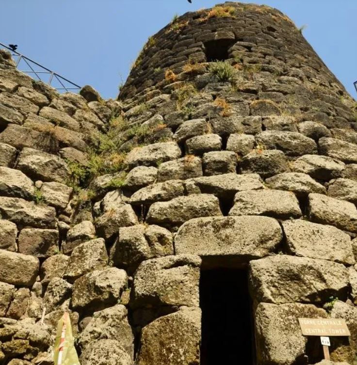 Una veduta dal basso di una torre in pietra consumata dal tempo, con un portale ad arco scuro e aperto alla base, sotto un cielo azzurro e limpido.