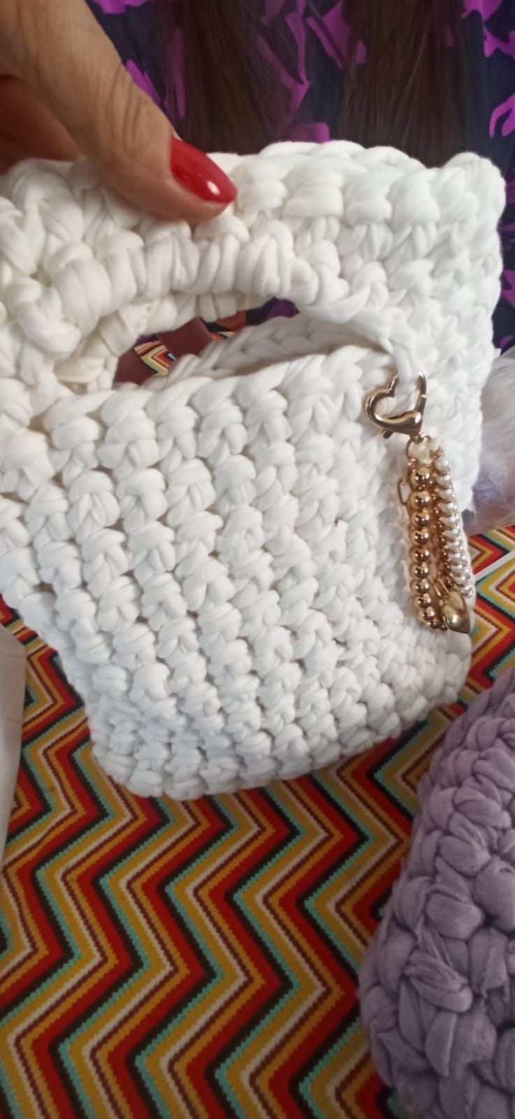 Bolso blanco de ganchillo con asa y borla dorada, sostenido por una mano con las uñas pintadas de rojo.