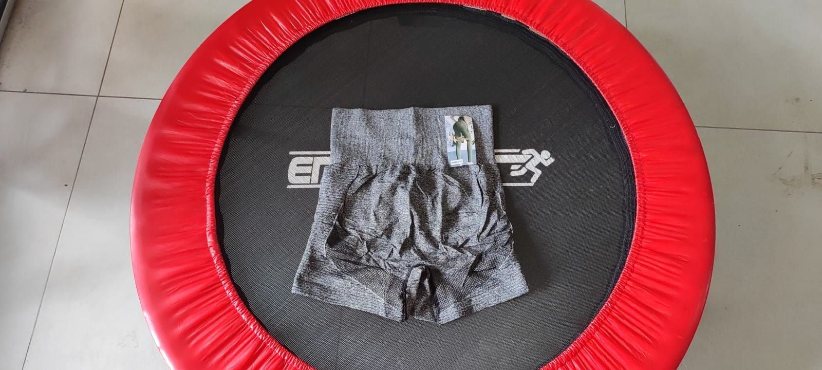 Pantalones cortos grises con etiqueta sobre una cama elástica negra con borde rojo.