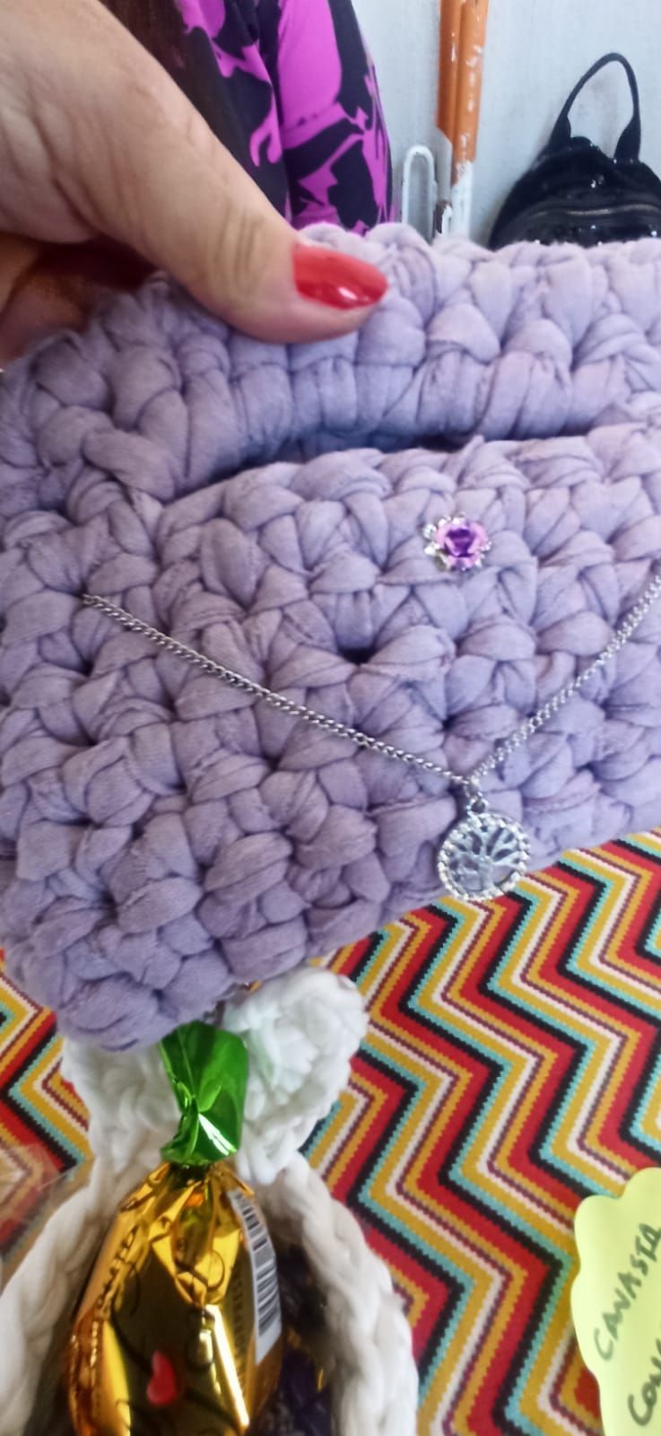 Una mano sostiene un objeto de ganchillo color lavanda adornado con un collar y una pequeña flor.
