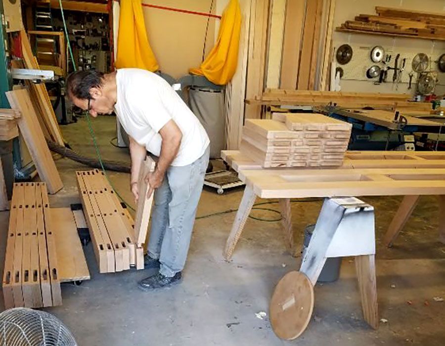 Woodworker bending over lumber in a cluttered workshop beside a workbench and stacked wood panels