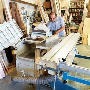Man working in a cluttered woodworking shop with a table saw and stacked lumber